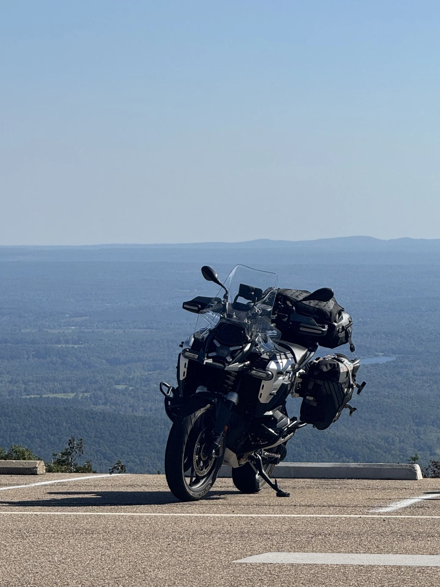 A motorcycle parked on the side of a mountain road overlooking a vast landscape of forested hills and valleys under a clear blue sky.