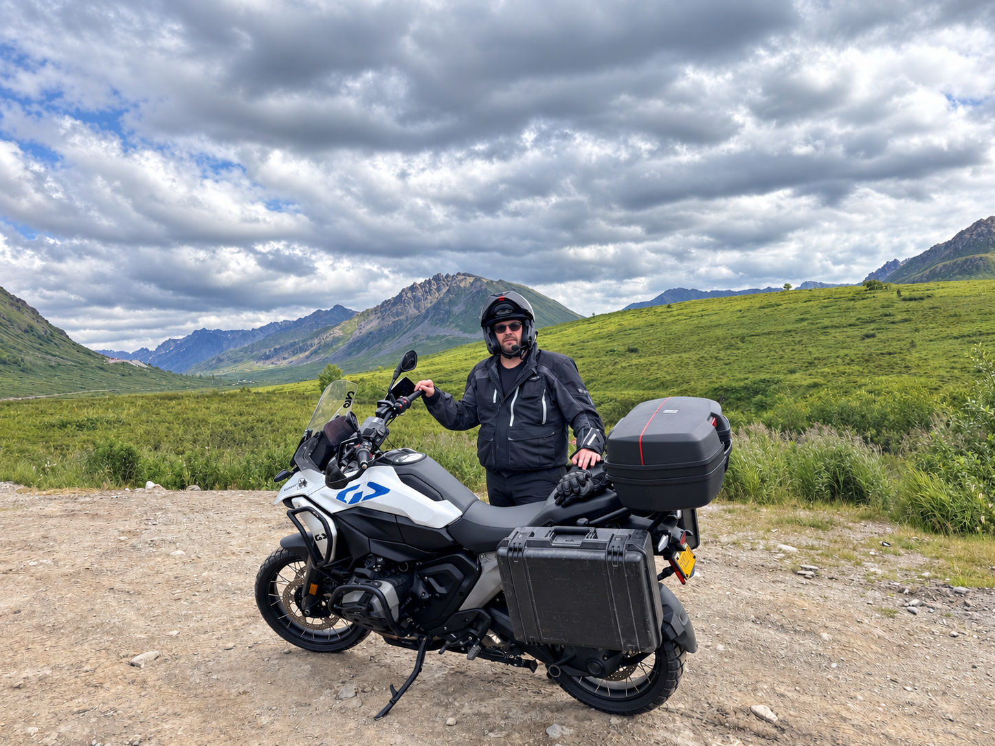 A man in black riding gear standing next to a black and white adventure motorcycle on a dirt path with green hills and mountains in the background under a cloudy sky.
