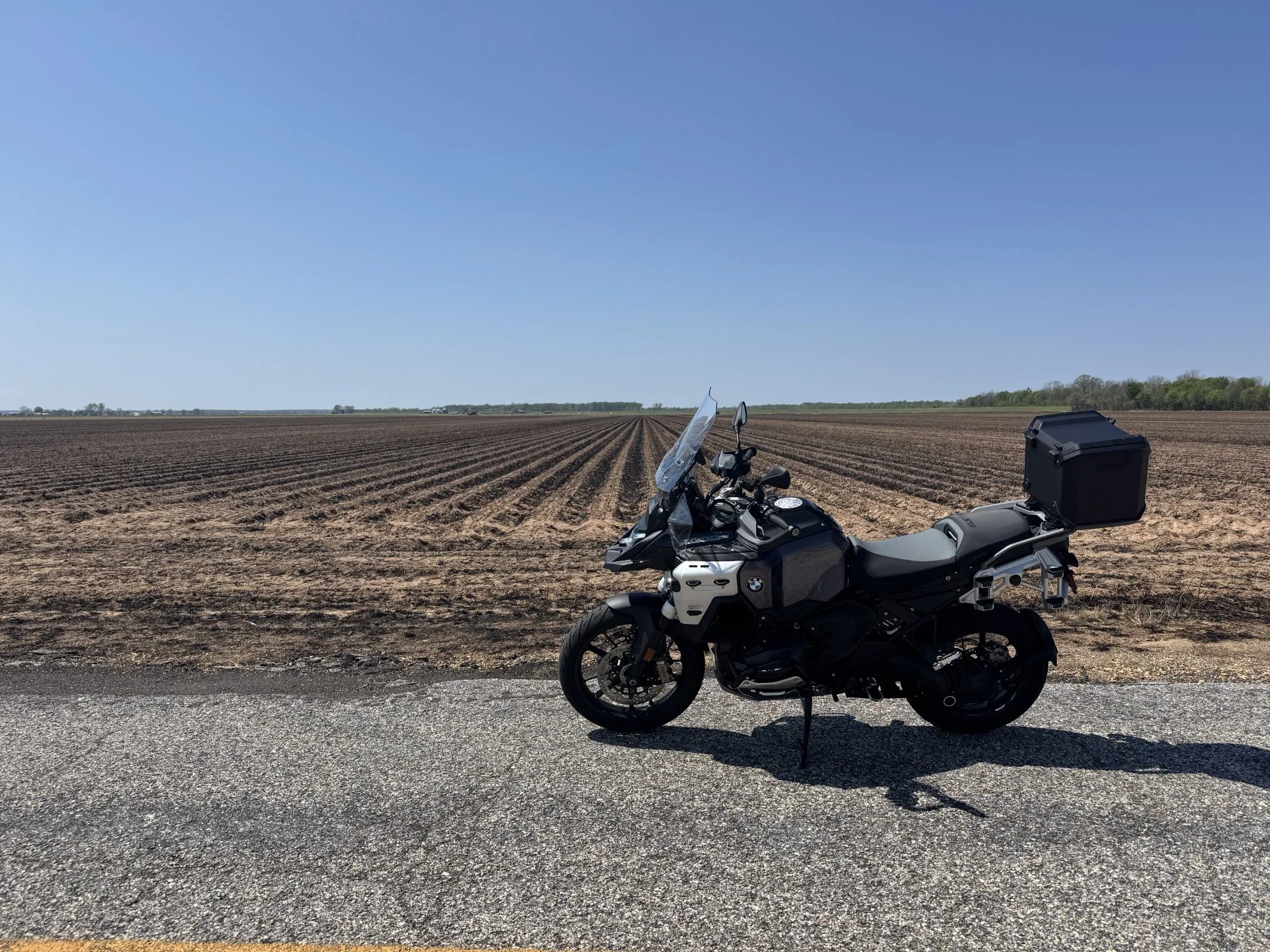 Black adventure motorcycle with luggage parked on side of road in front of plowed farmland under clear blue sky