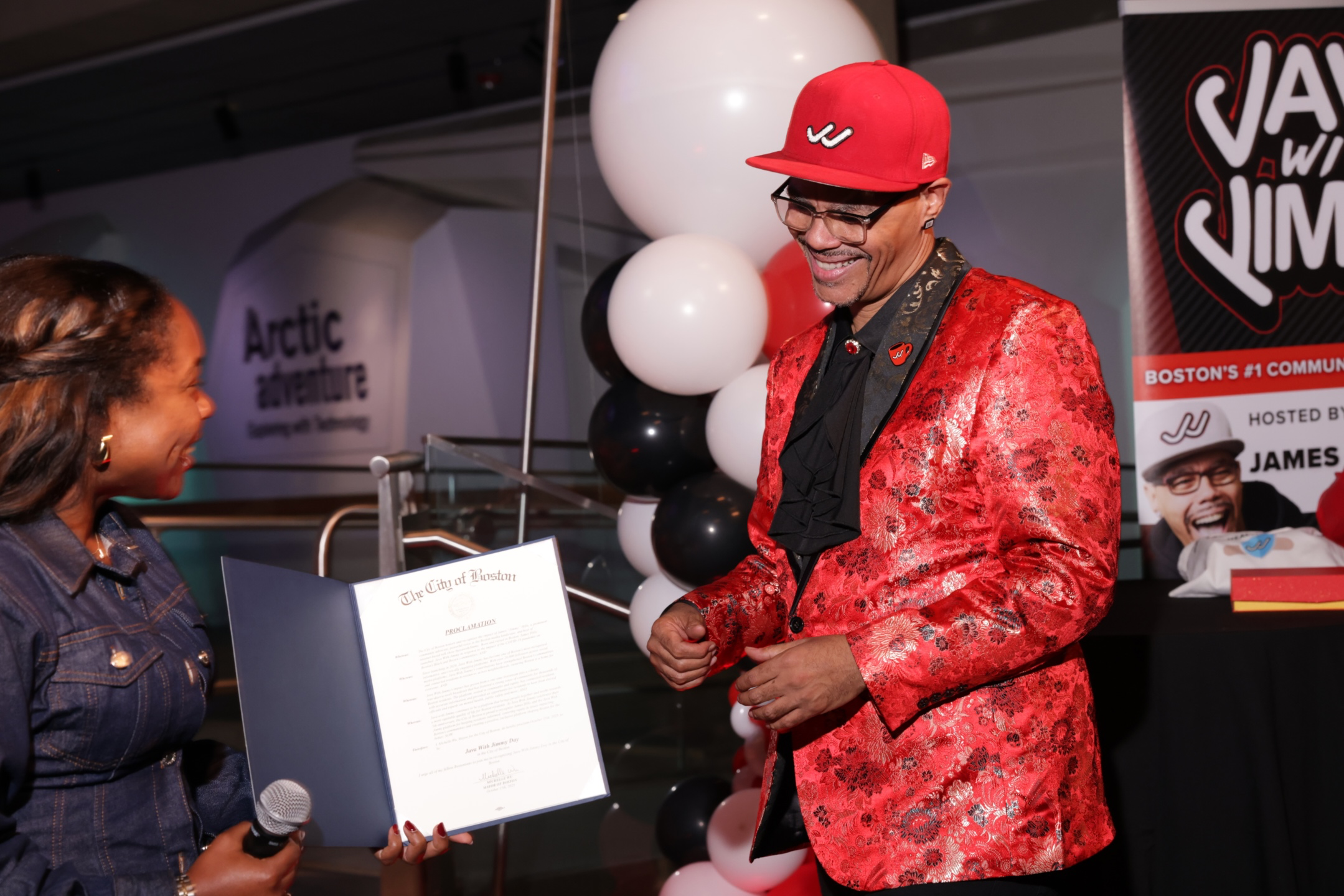 A man wearing a red hat, glasses, and a red patterned blazer is smiling and talking to a woman holding a microphone and a framed document at an event. There are white and black balloons in the background and a banner with text.
