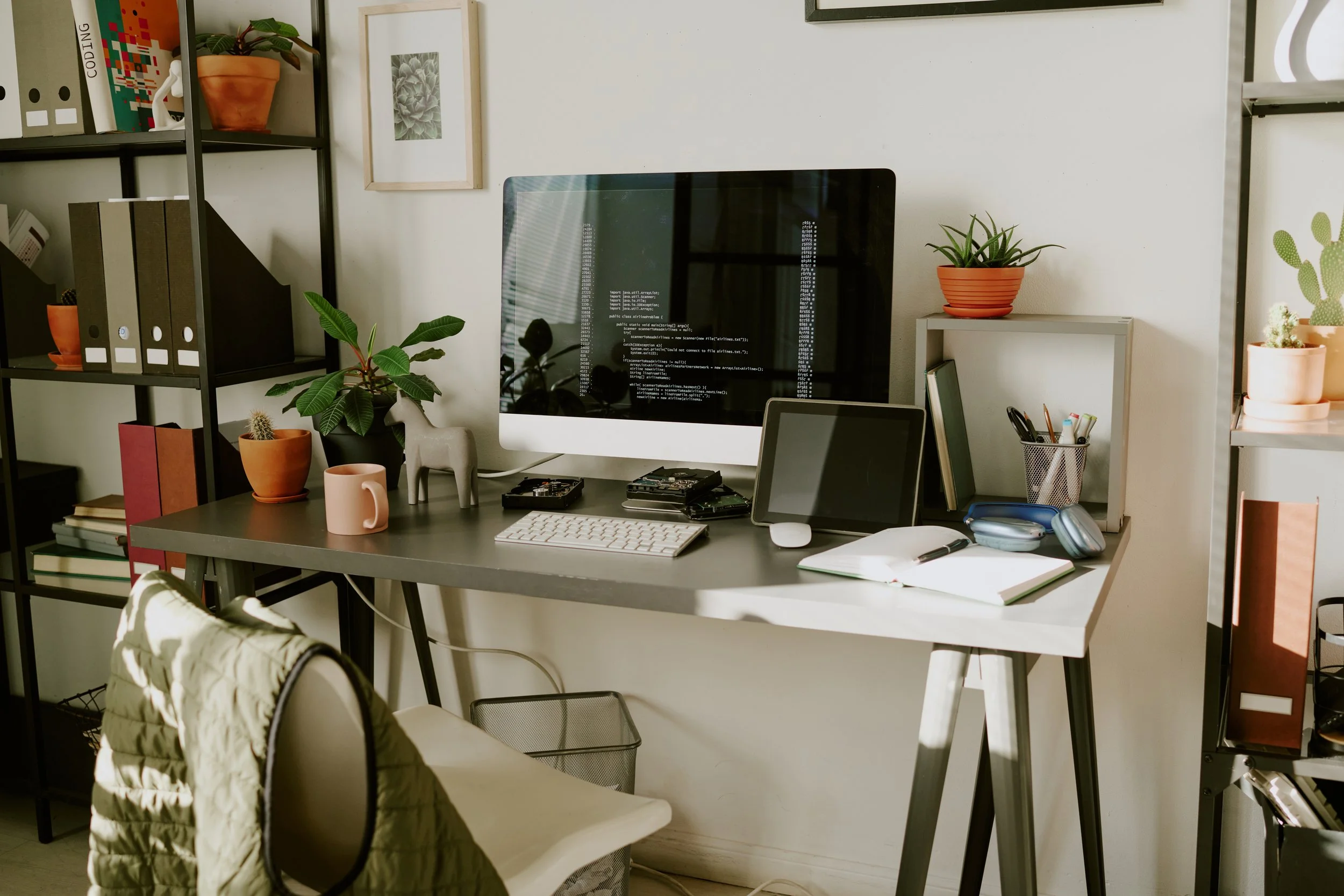 Home office workspace with a computer monitor displaying coding scripts, a keyboard, mouse, notebooks, headphones, and various potted plants on shelves and desks, with a person sitting in front of it.