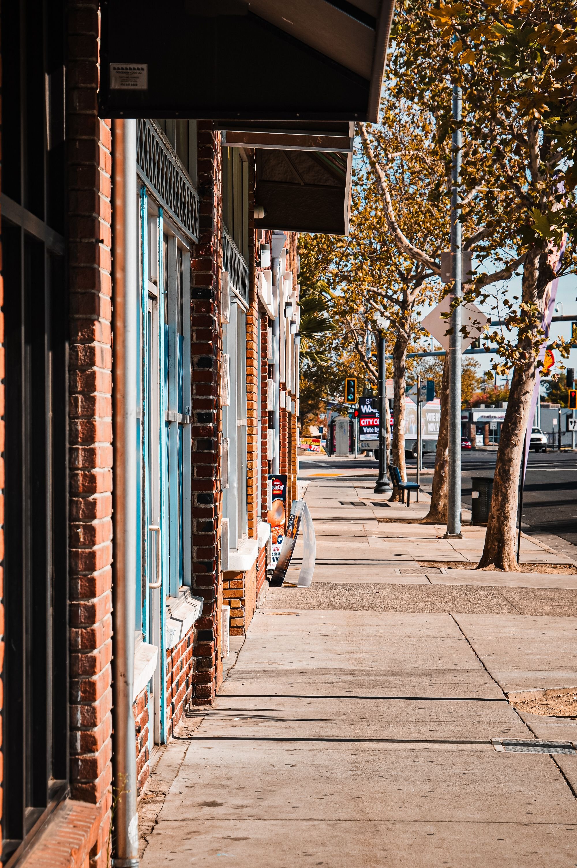 Empty sidewalk with storefronts on the left, trees with autumn leaves on the right, and street signs and traffic lights in the background.
