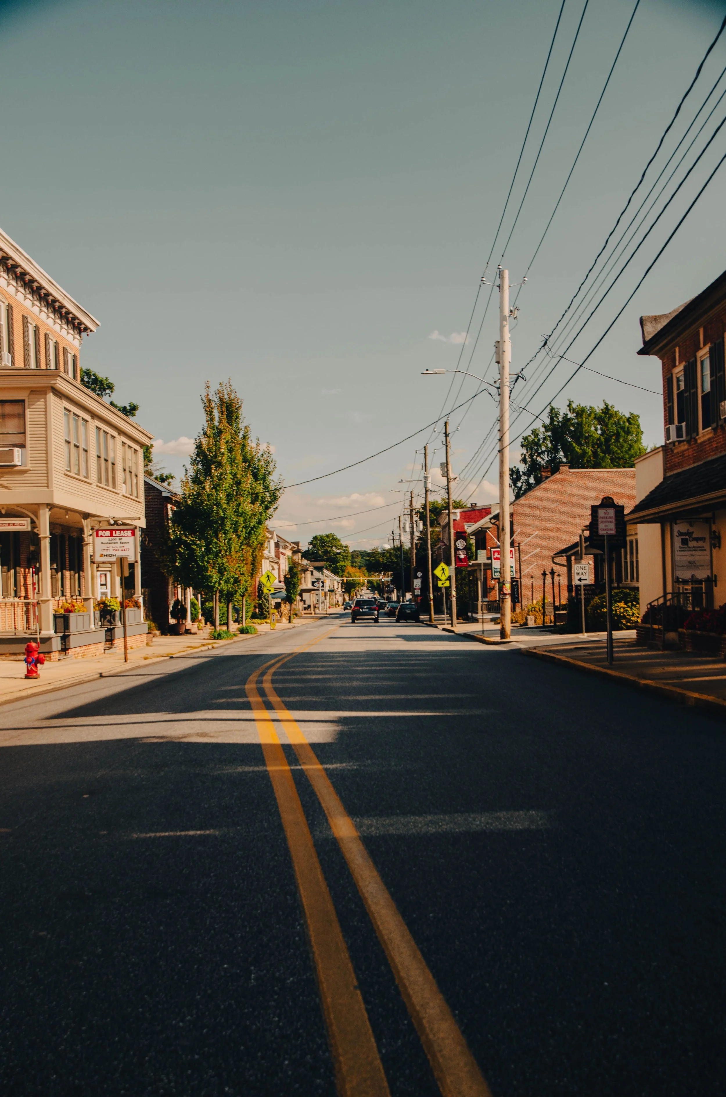 A quiet street in a small town with houses and businesses, power lines overhead, and a few cars parked along the curb, under a clear sky.