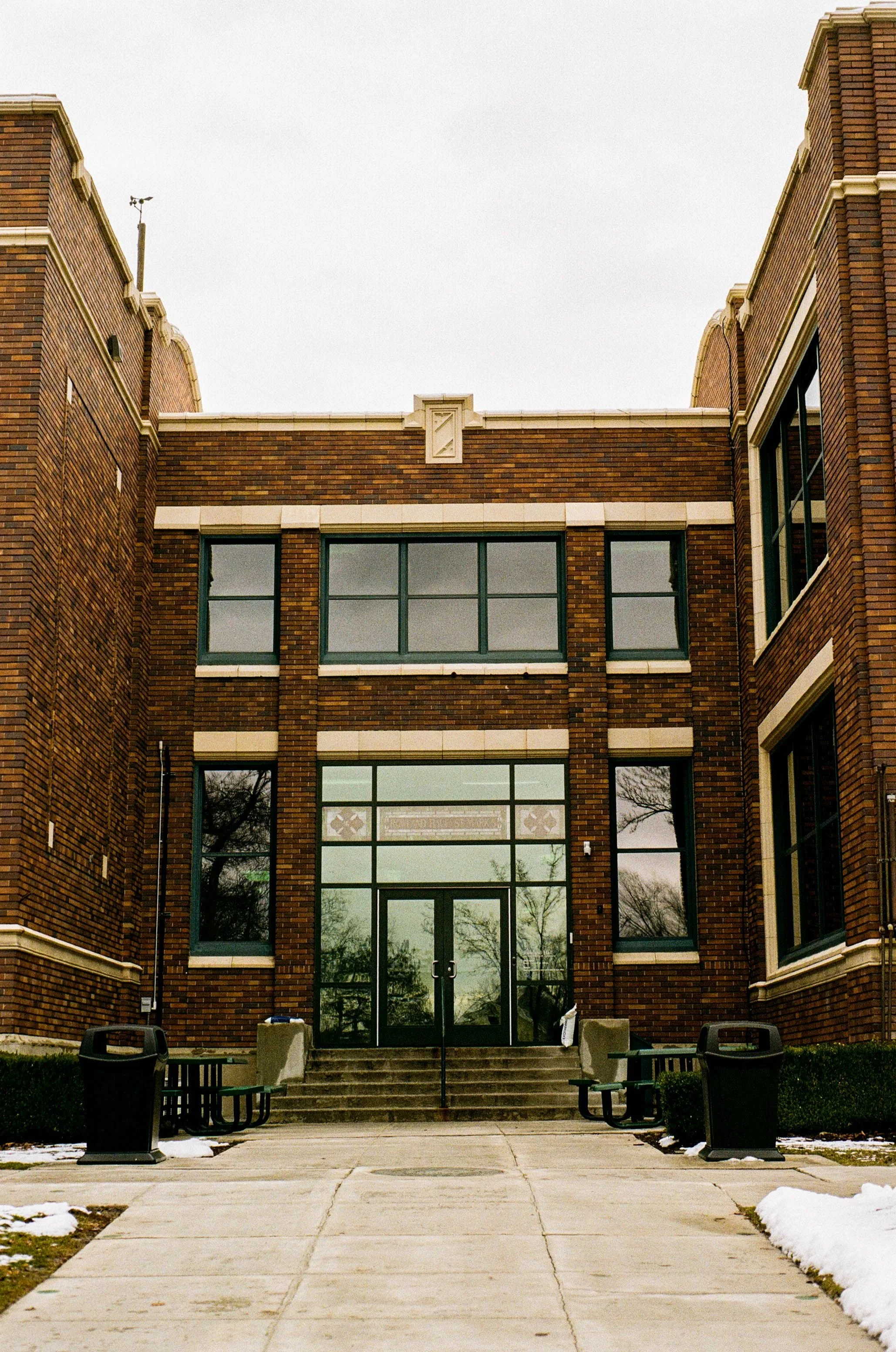 Brick building with large glass doors and windows, concrete stairs, benches, trash cans, and some patches of snow on the ground.