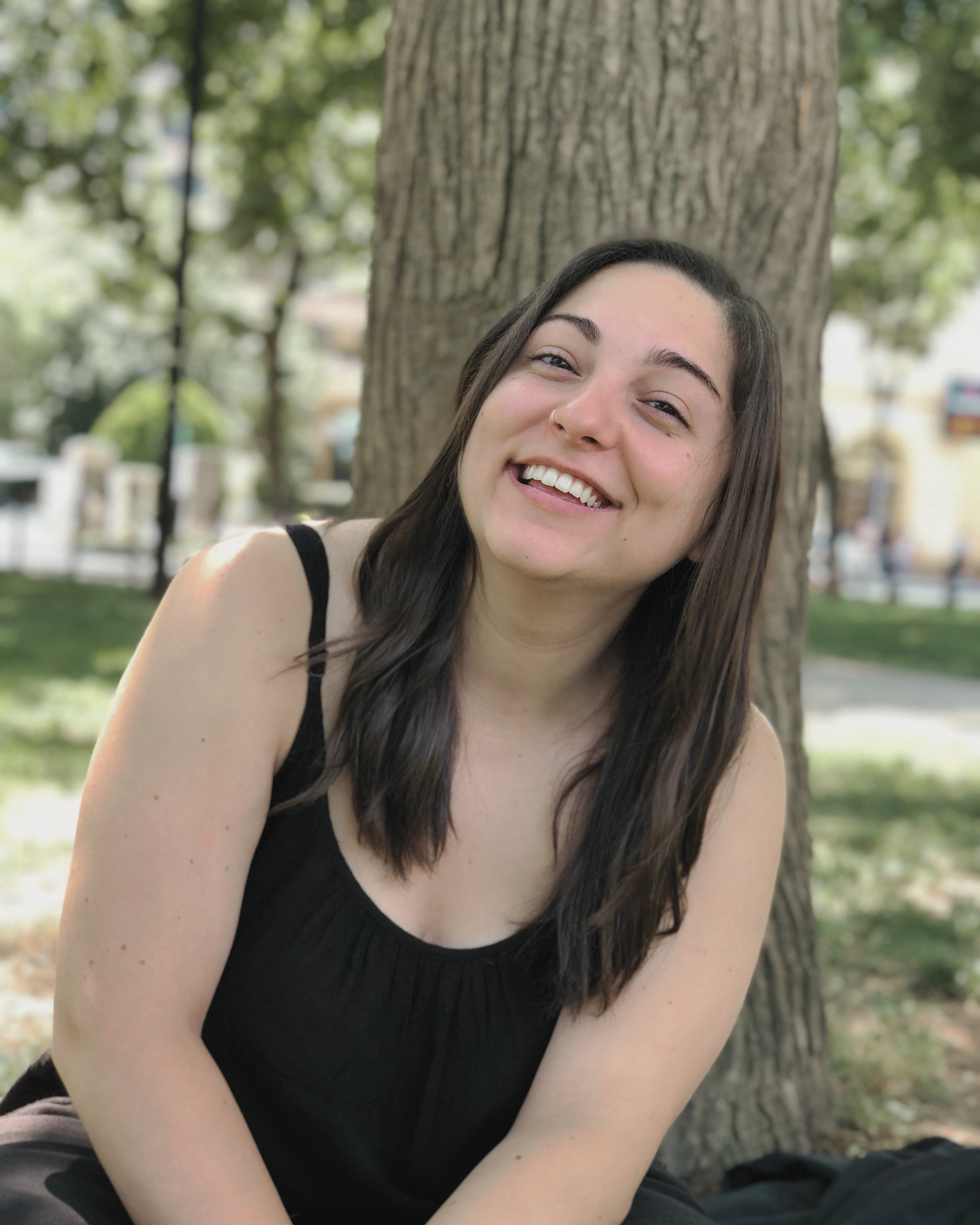 A young woman with dark hair and a small nose ring smiling and leaning against a tree outdoors during daytime, with green leafy trees in the background.