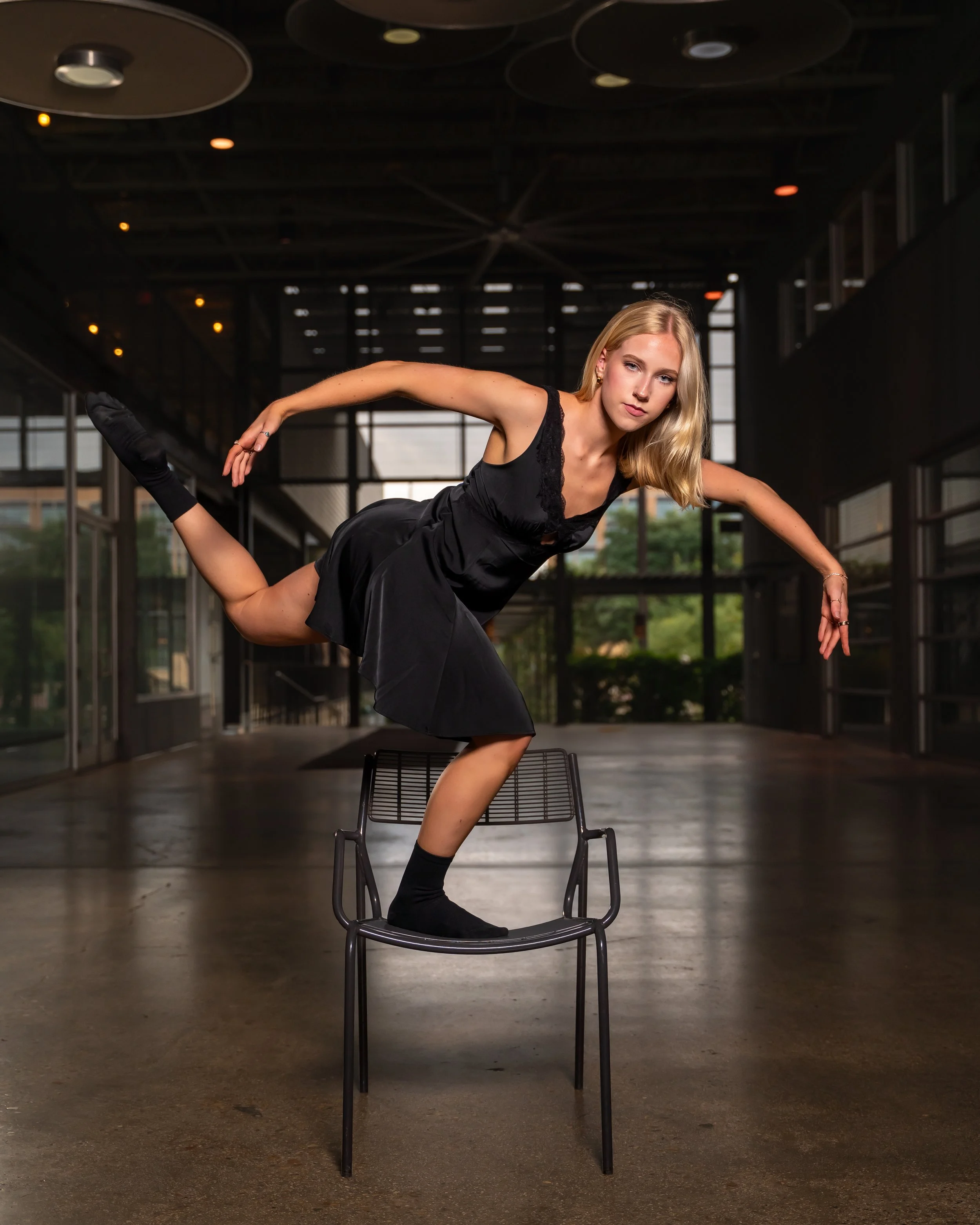 A woman in black dance attire is balancing on a chair, with one leg bent behind her and one arm extended forward and the other arm extended backward, in an indoor space with modern architecture and large glass windows.