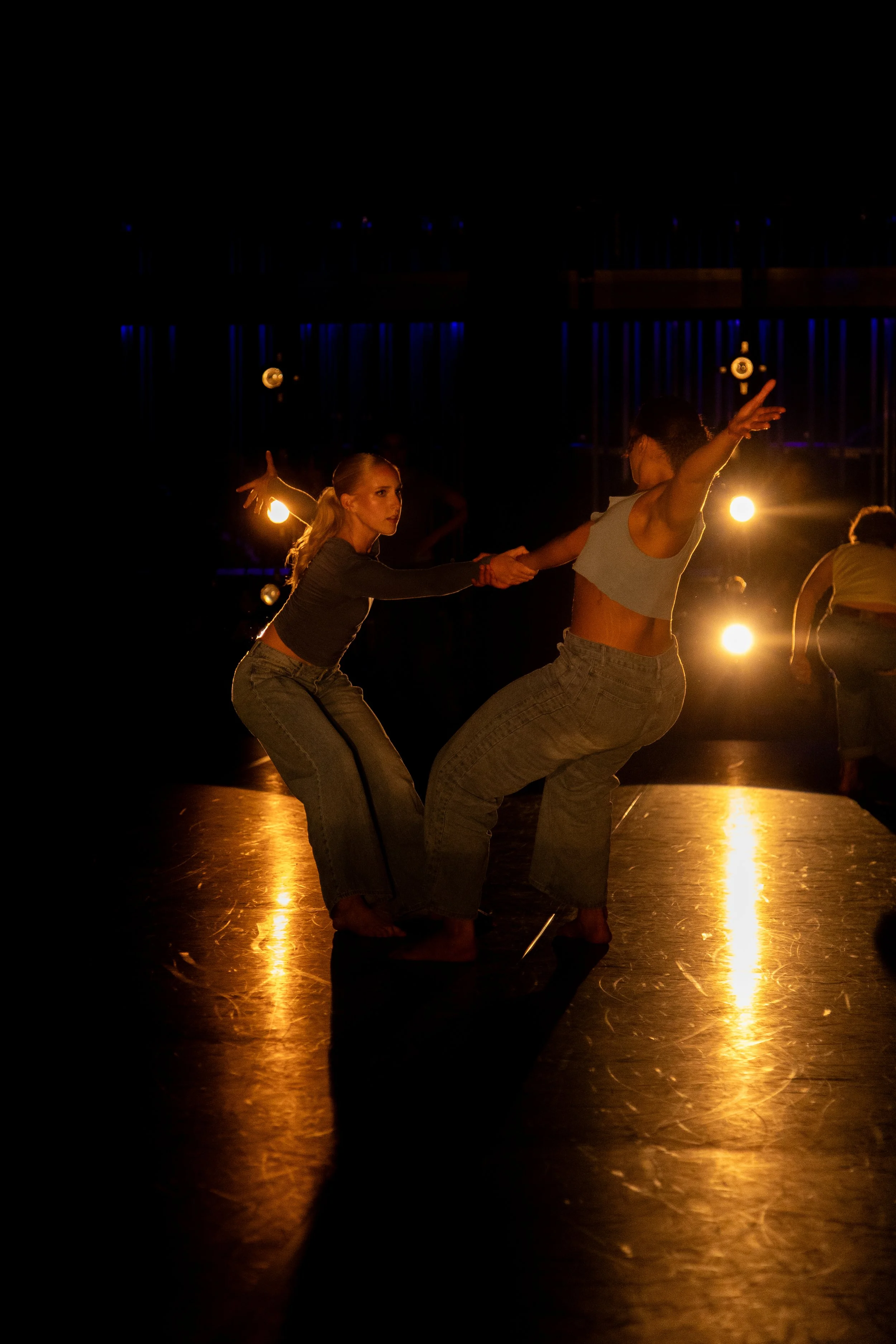Two dancers are performing on stage under warm lighting, holding hands and leaning back as part of their dance routine.