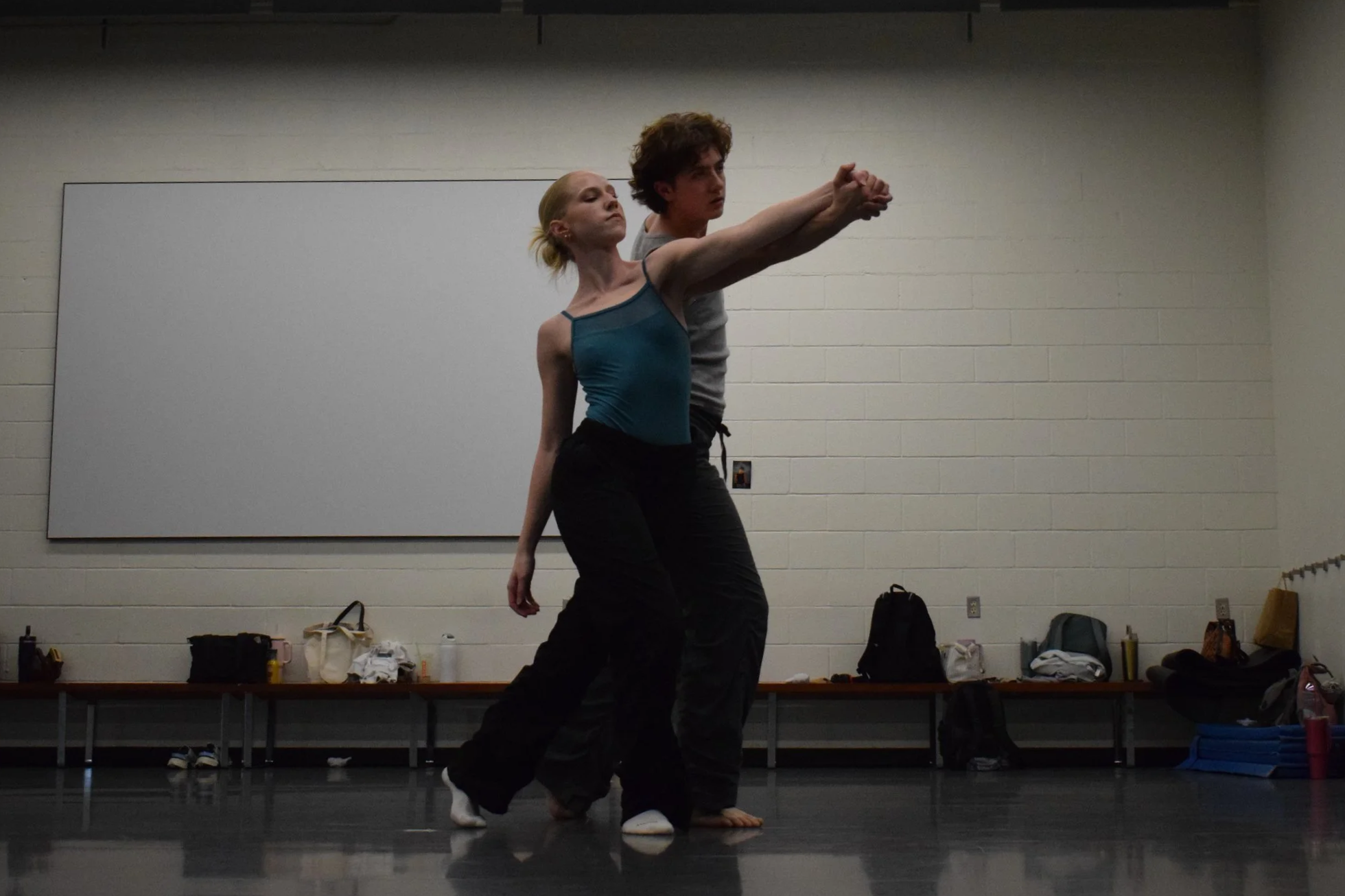 Two dancers practicing together in a dance studio, one male and one female, with a whiteboard and bags on a shelf behind them.