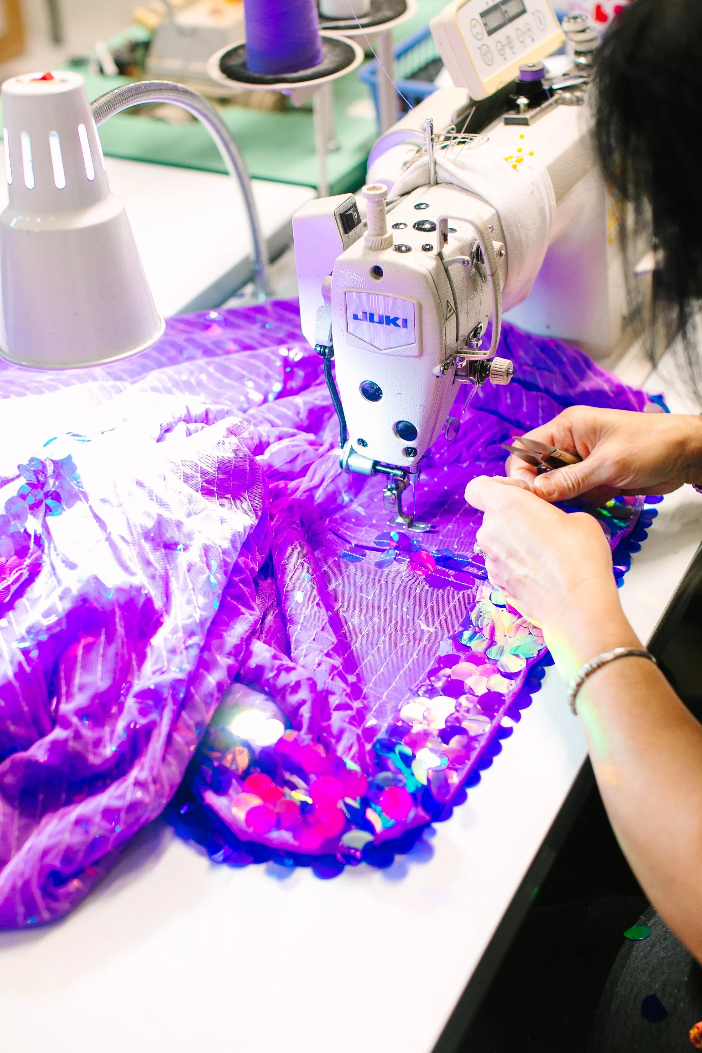 Close-up of a sewing machine stitching colorful purple and pink fabric with sequins, with a person's hands guiding the fabric.