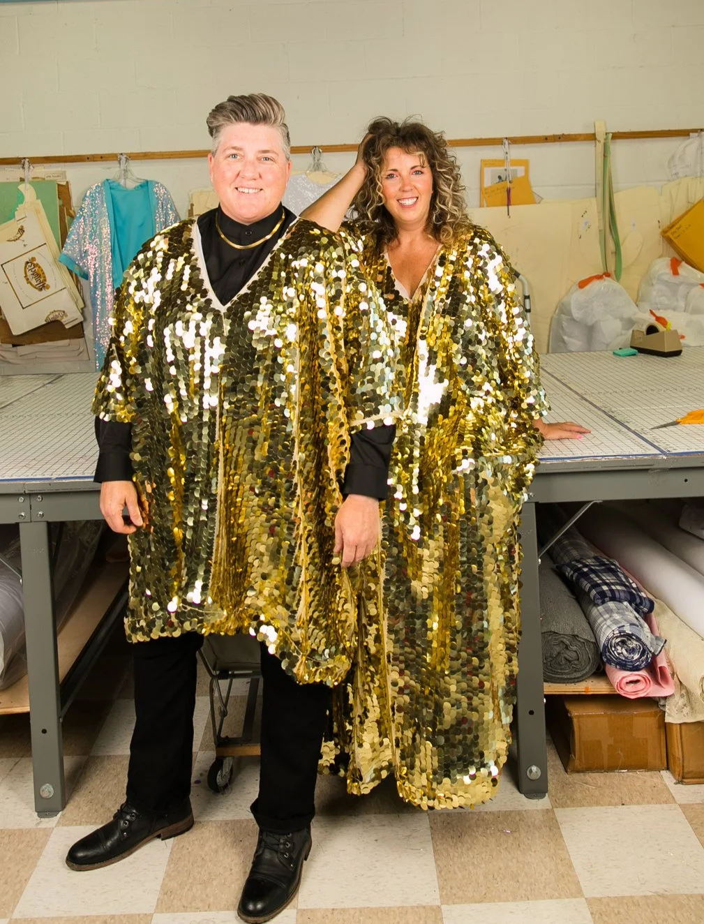 Two people standing side by side wearing matching gold sequin coats in a workroom. The man is smiling with white hair, dressed in black pants and shoes, with a black shirt and a gold necklace. The woman has curly hair, smiling, with her hand on her head, wearing black pants. Behind them is a worktable with patterns, fabrics, and sewing supplies.