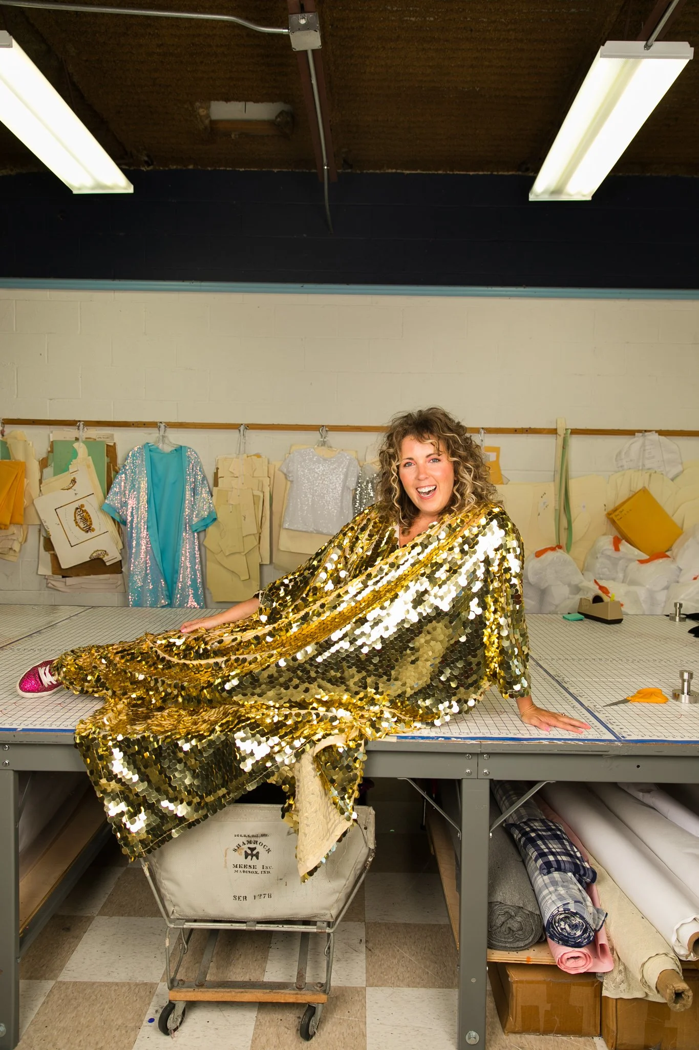 A woman dressed in a shiny gold sequin dress sitting on a large cutting table in a sewing room. Behind her are fabric swatches and sewing patterns hanging on the wall, with rolls of fabric and sewing supplies stored below the table.