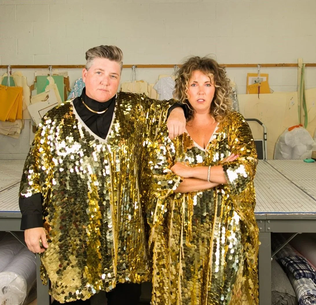 Two people standing side by side wearing shiny gold sequin outfits in a workspace with mannequin patterns in the background.
