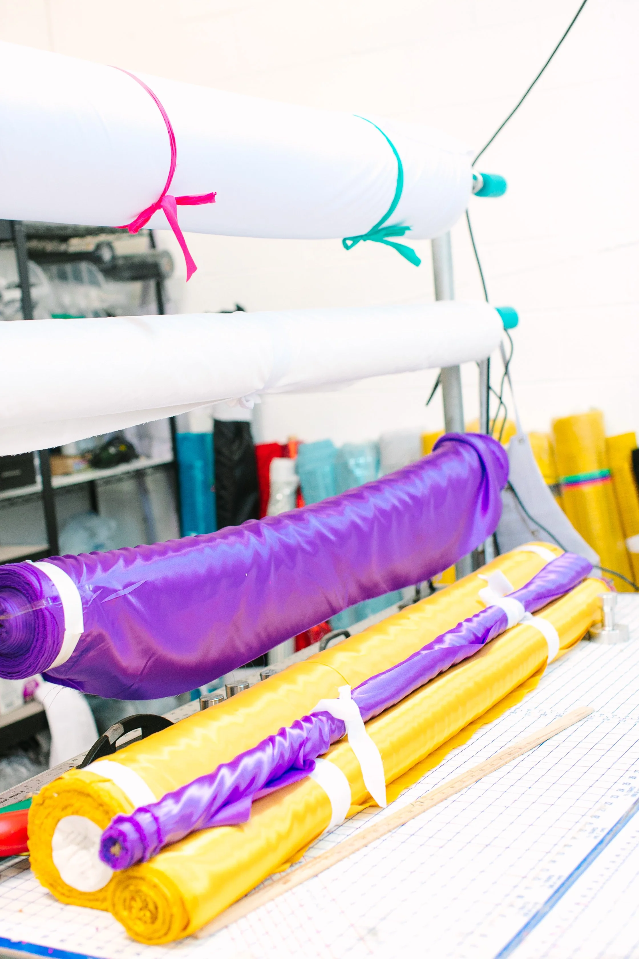 Rolls of yellow and purple fabric or material on a work table with a grid pattern, surrounded by tools and supplies in a workshop or sewing room.