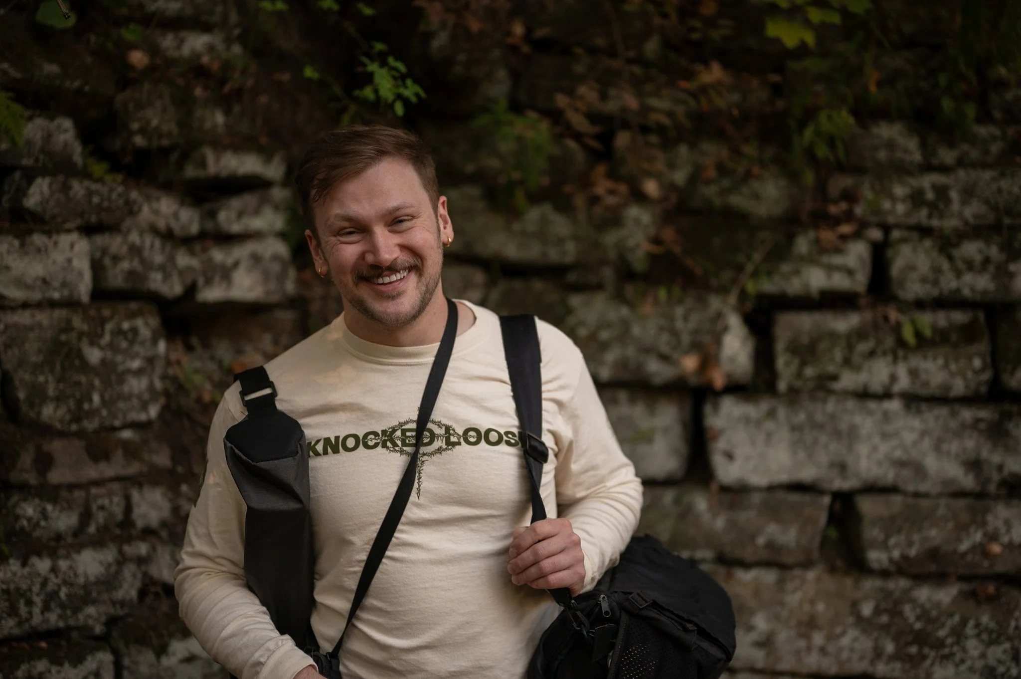Portrait of Sterling Dunham with short brown hair, beard, and earrings, smiling and wearing a beige long-sleeve shirt with 'KNOCKED LOOSE' logo, carrying multiple camera bags, standing outdoors against a stone wall with greenery.