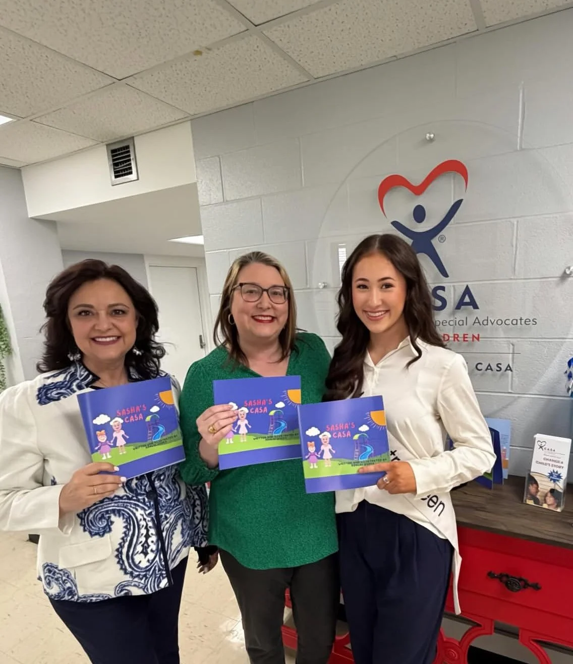Three women standing together indoors, each holding a colorful booklet titled 'Sasha's Casa,' smiling at the camera.