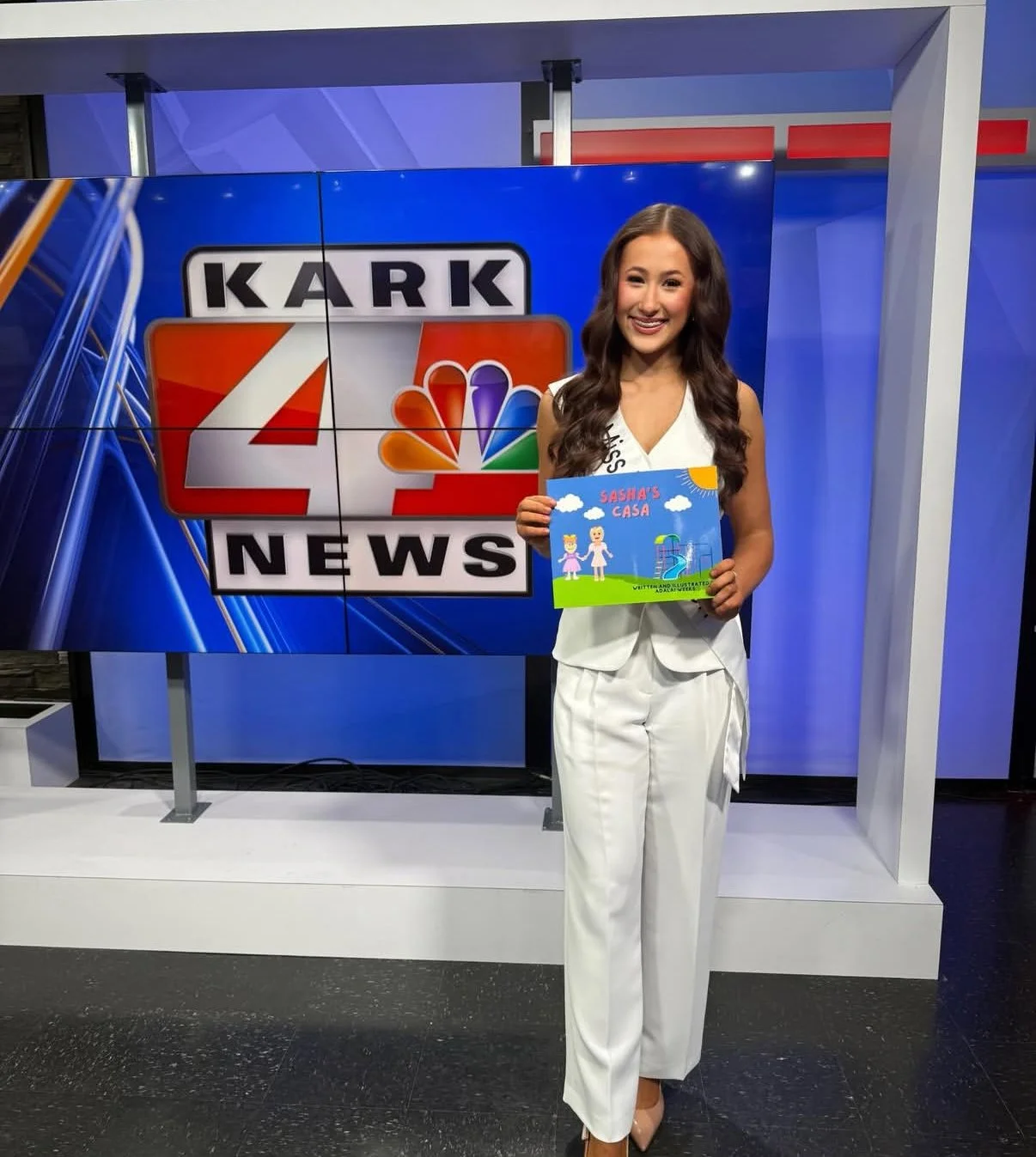 A woman in a white suit smiling while holding a children's drawing titled 'Sasha's Casa' in front of a television studio background with news station graphics.