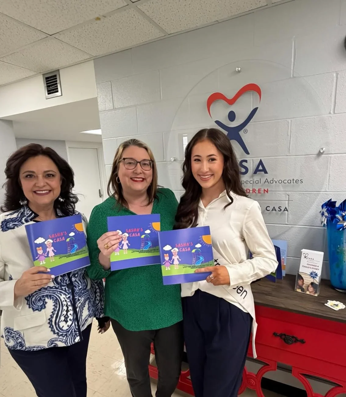 Three women smiling and holding children's books titled 'Sasha's Casa' inside an office or lobby with a CASA logo on the wall behind them.