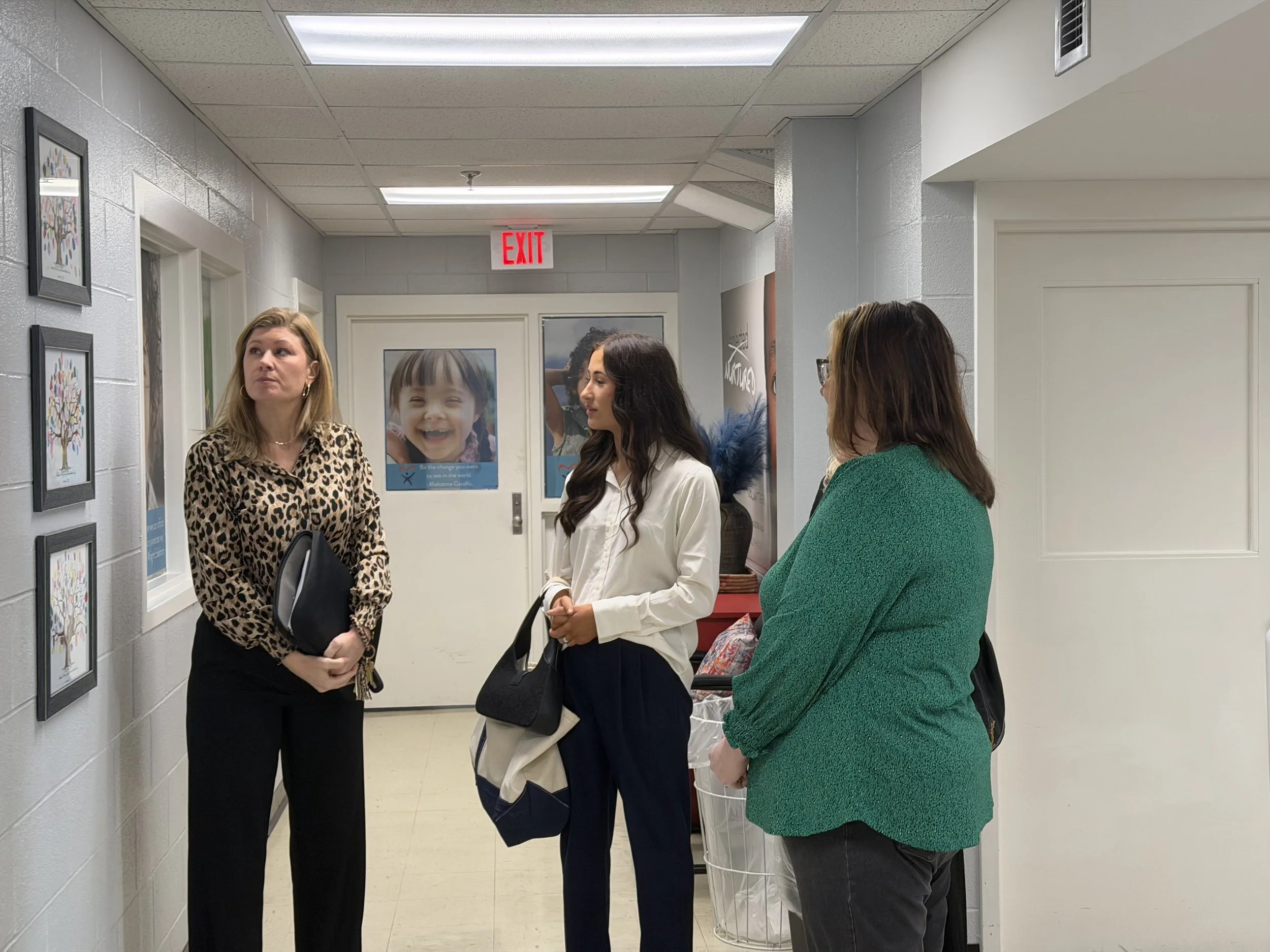 Three women standing and talking in a hallway with artwork and posters on the walls, including an image of a smiling child.