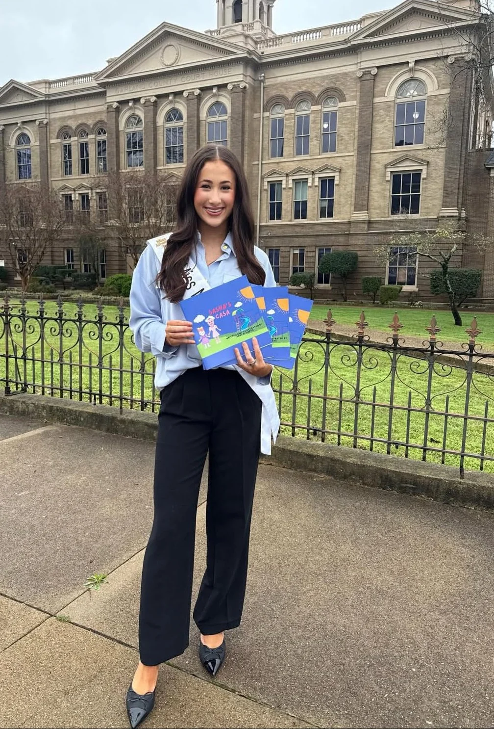 A young woman dressed in business attire is standing outdoors in front of a historic courthouse building, smiling and holding four colorful booklets titled 'Basha's Casa' featuring illustrations of children, clouds, and a fountain.