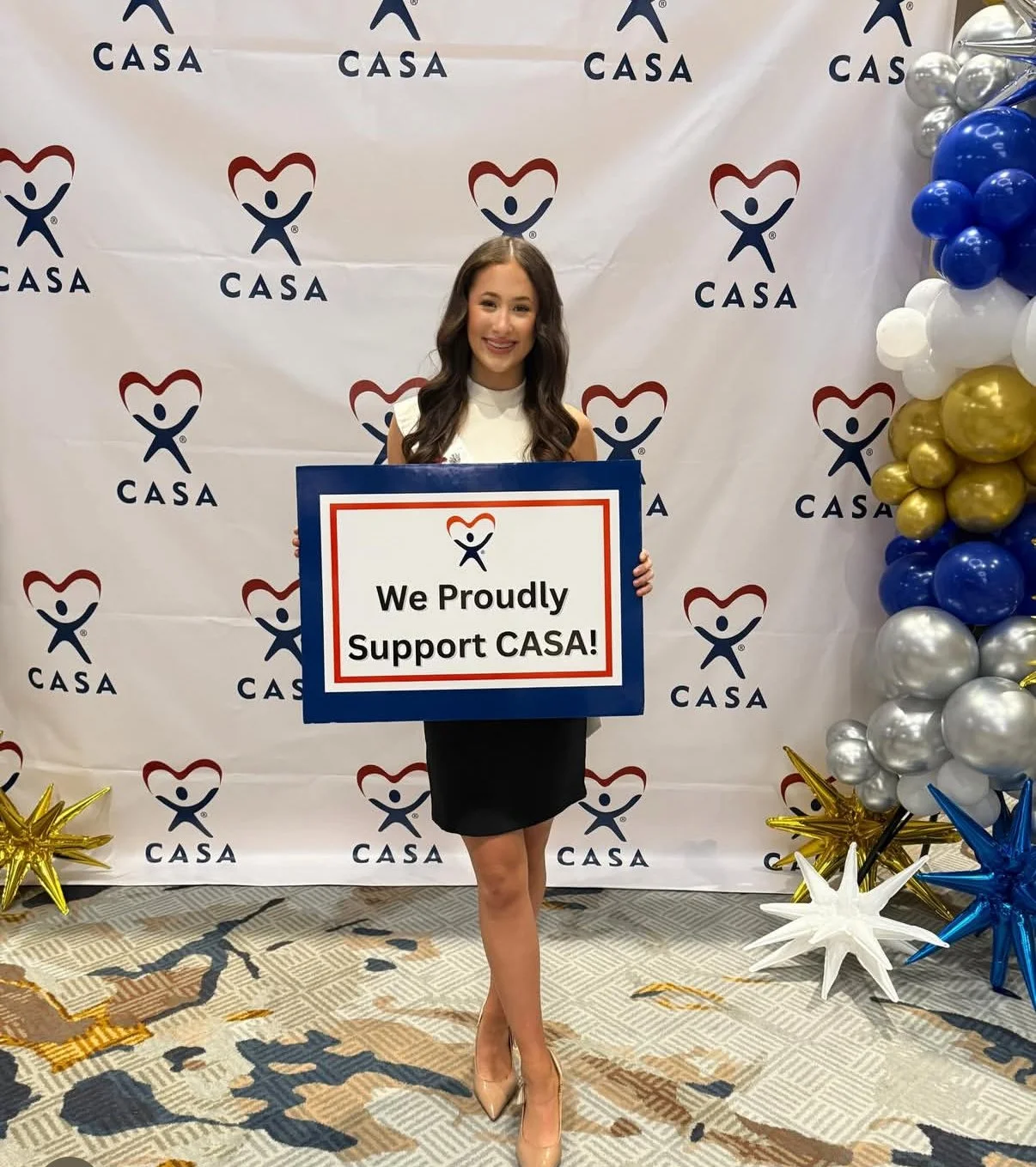 A young woman standing indoors at a CASA support event, holding a sign that says 'We Proudly Support CASA!' in front of a backdrop with the CASA logo. There are balloons in gold, silver, blue, white, and star-shaped decorations on the floor.