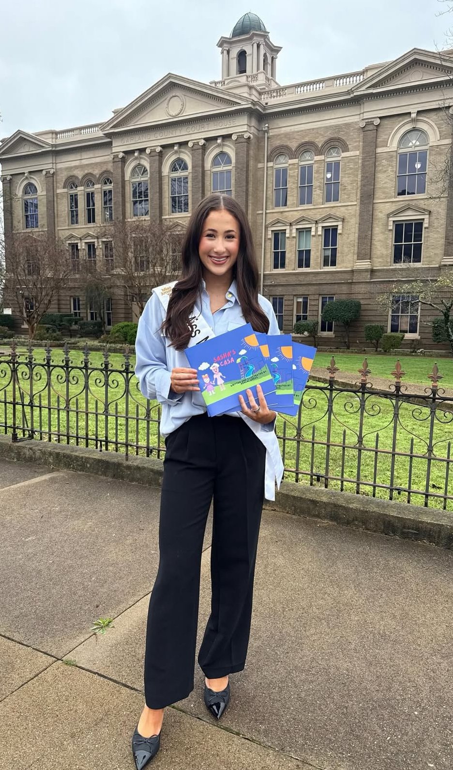 A young woman standing outside in front of a historic courthouse, holding pamphlets for a program called 'Sahas Casa,' smiling at the camera, dressed in professional attire.