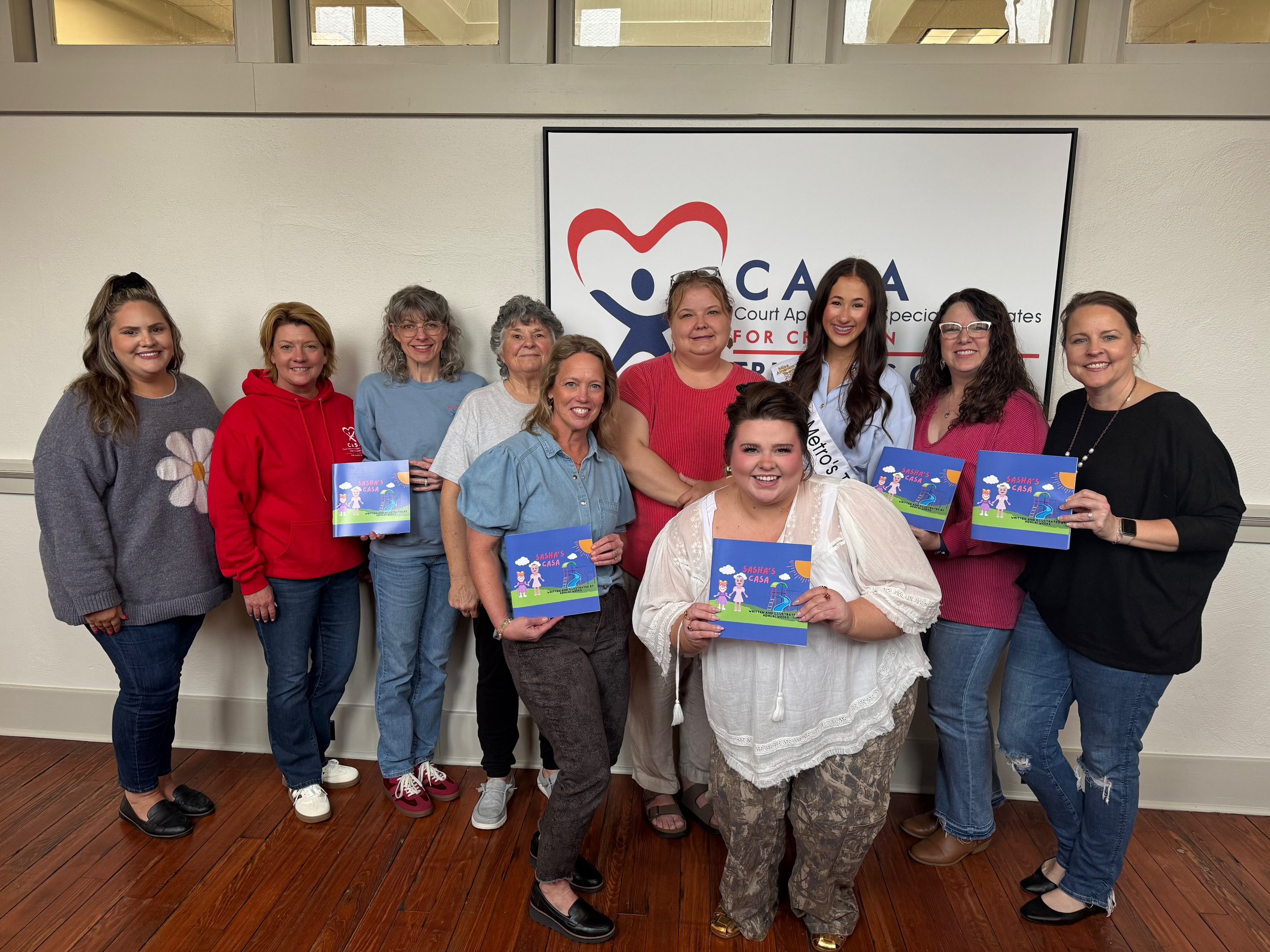Group of women standing in front of a sign that reads "Court Appointed Special Advocates for Children" with a logo of a person inside a heart. Several women are holding colorful children's books titled "Sasha's Casa." The women are smiling and dressed casually.