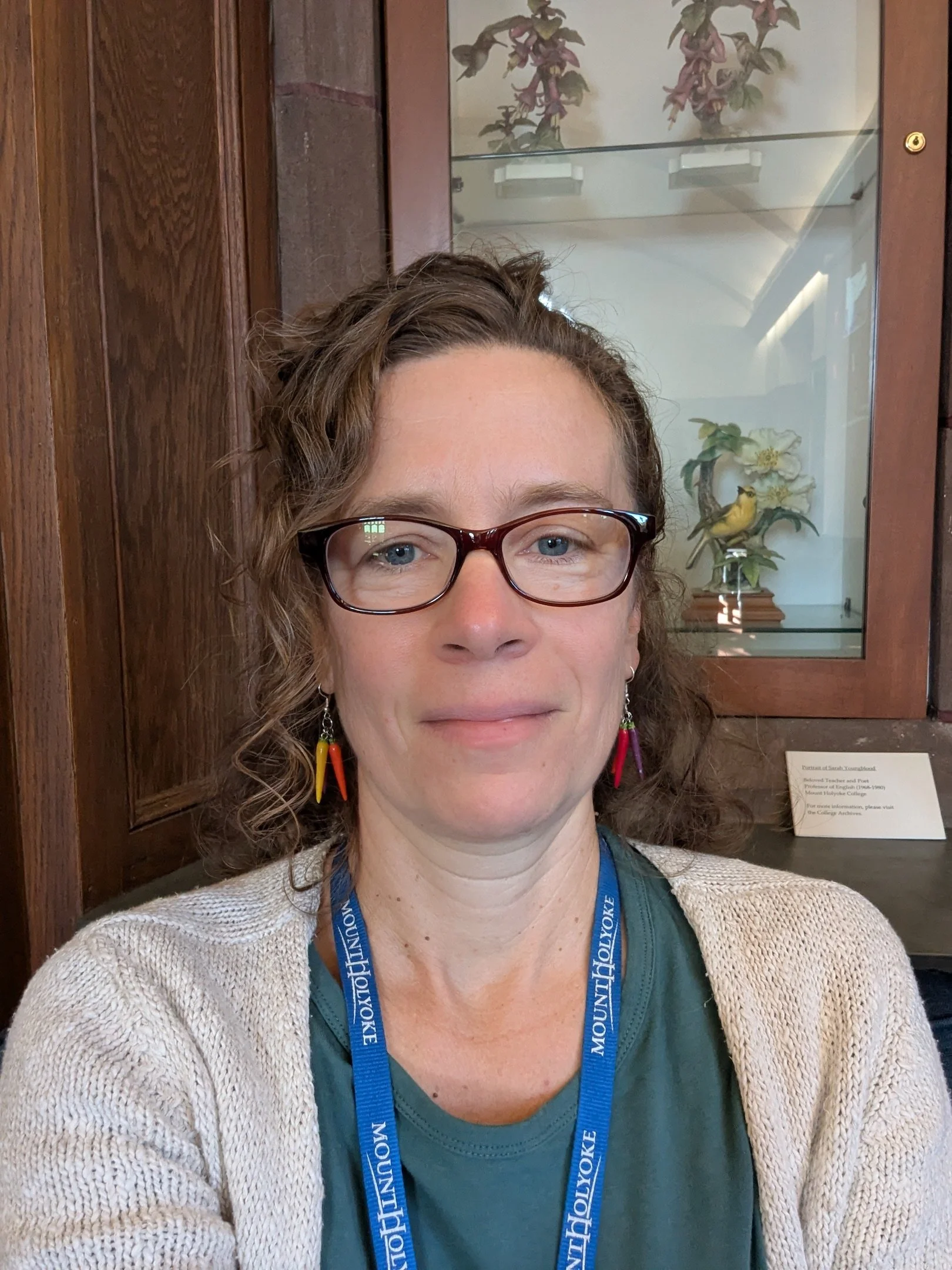 Close-up of a woman with glasses and earrings, smiling, standing in front of a wooden display case.