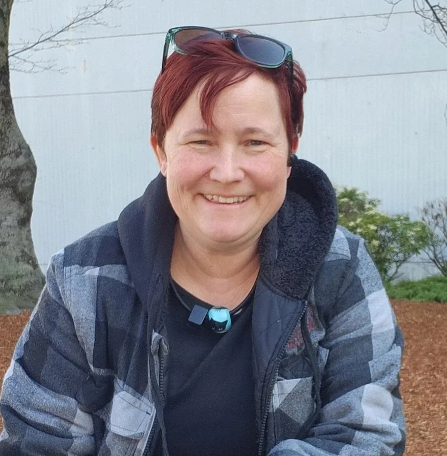 A woman with short red hair smiling outdoors, wearing sunglasses on her head, and a black shirt with a plaid jacket.