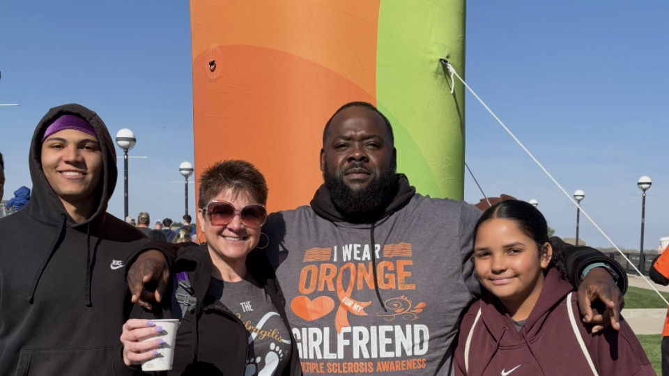 Group of four people smiling and posing for a photo outdoors near a tall, colorful, orange and green balloon, with a clear blue sky in the background.