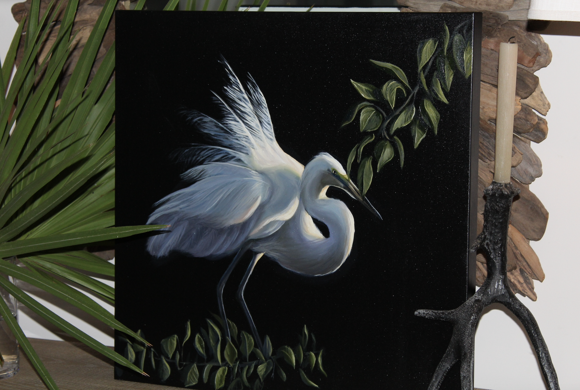 Painting of a white heron with a long beak, surrounded by green leaves, on a black background.