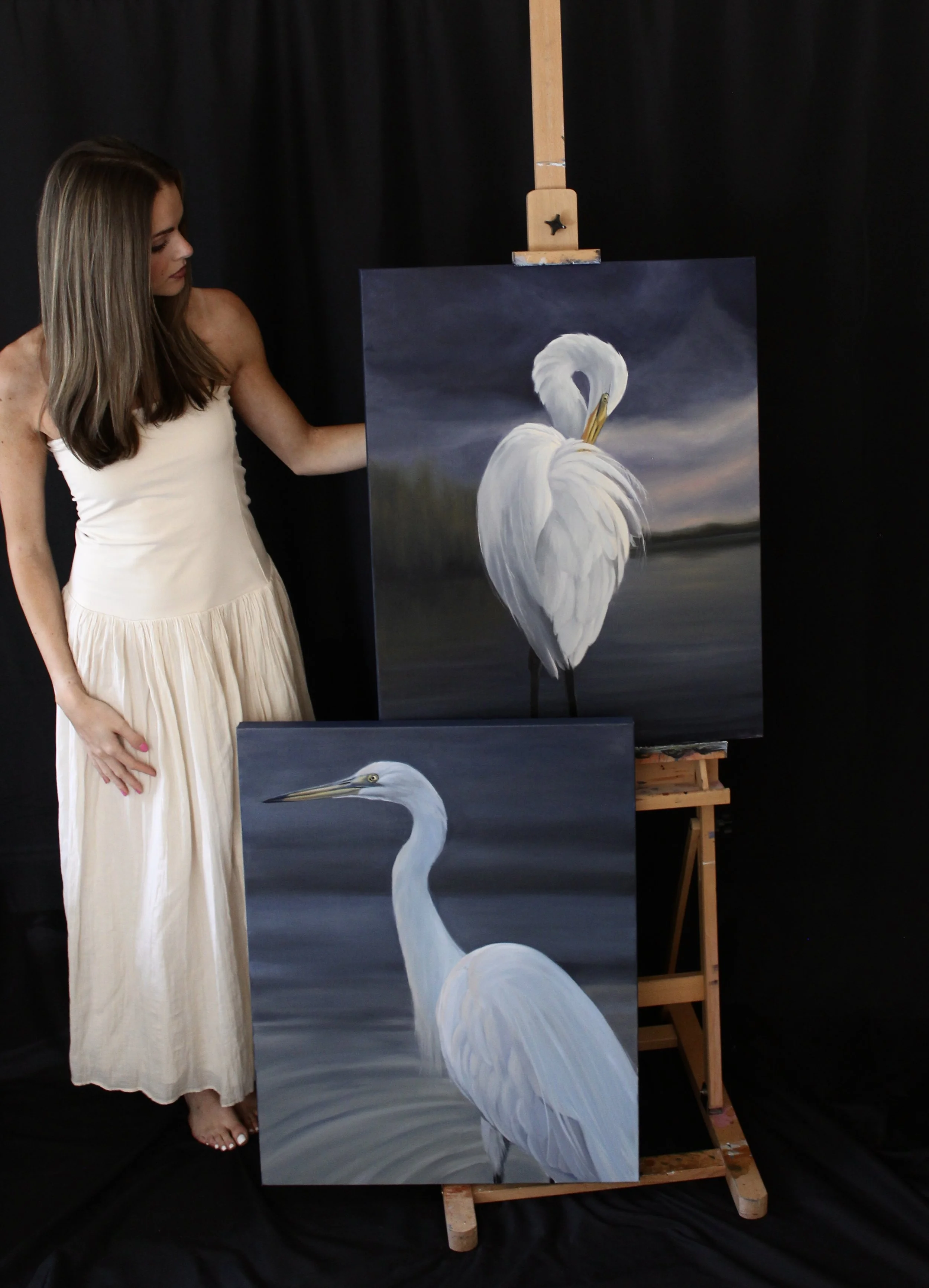 A woman in a white dress is standing beside a painting of a white heron, which is on an easel. The background is black, and the painting depicts the bird with a dark, cloudy sky.