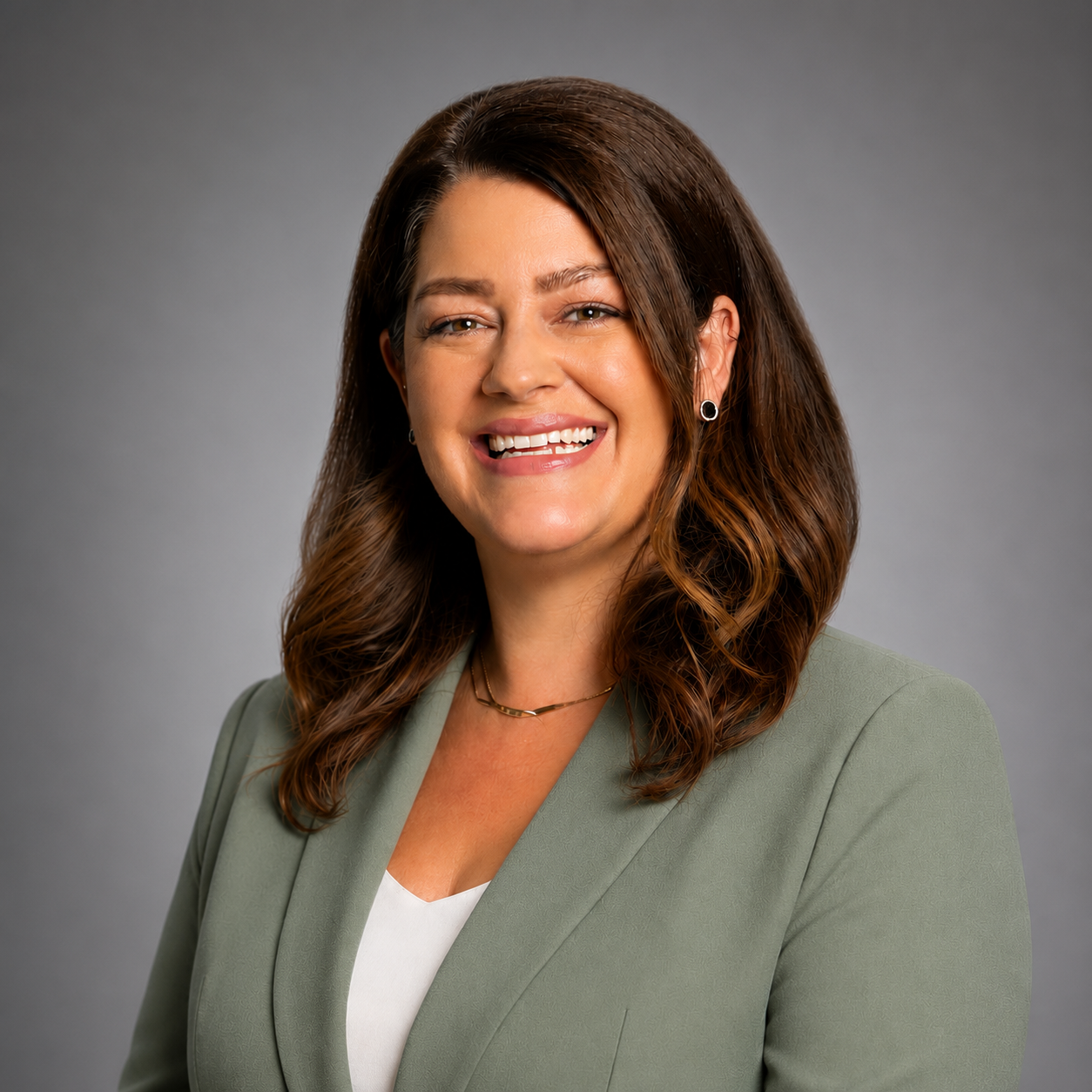 Professional woman with shoulder-length wavy brown hair, smiling, wearing a light green blazer, white top, and jewelry, against a neutral gray background.