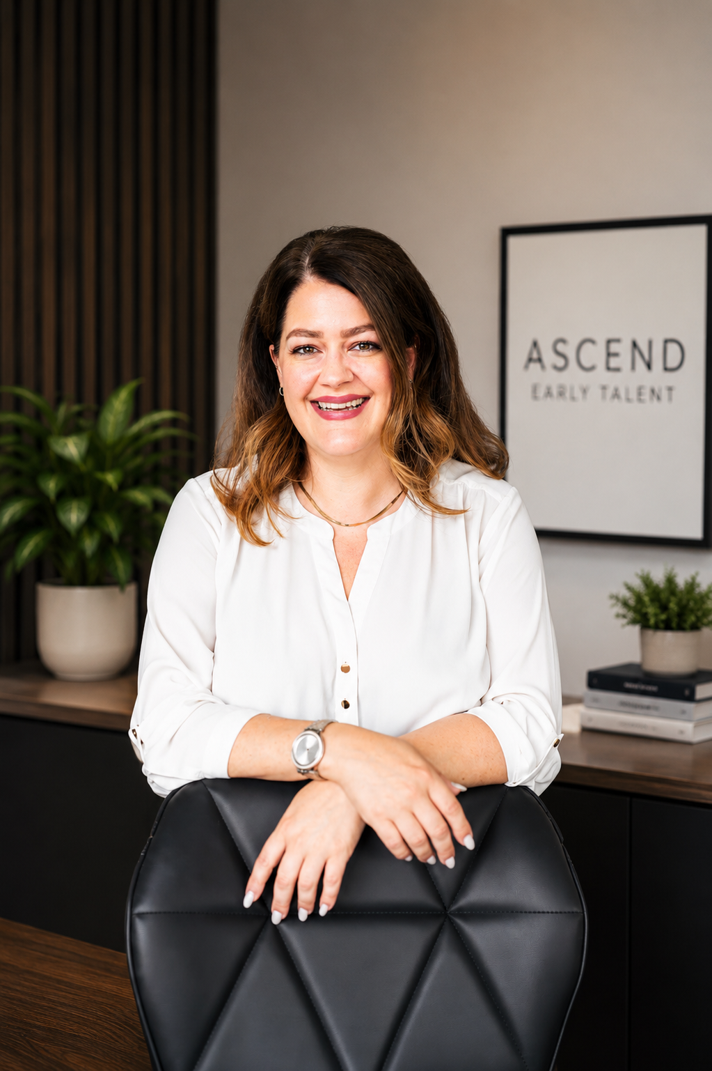 A woman with shoulder-length brown hair smiling, wearing a white blouse, sitting behind a black office chair with her arms resting on the back. In the background, a sign says 'ASCEND EARLY TALENT' with a potted plant and some books on a wooden desk.