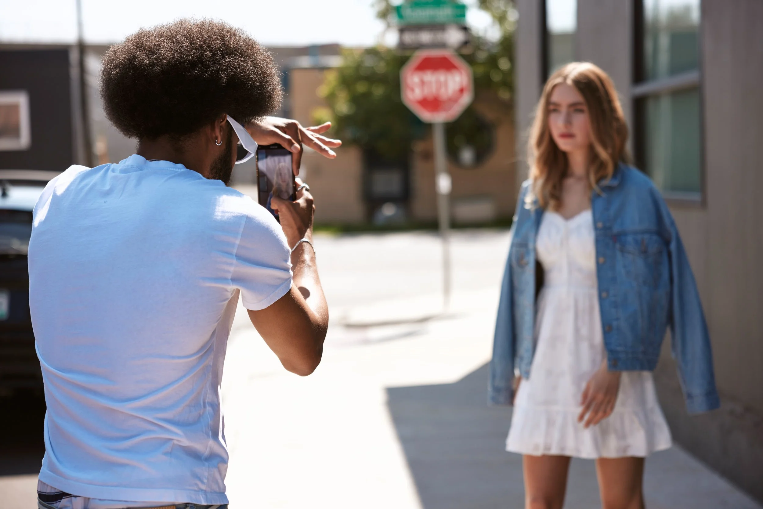 A man with curly hair taking a photograph of a woman with brown hair standing outdoors on a city street.