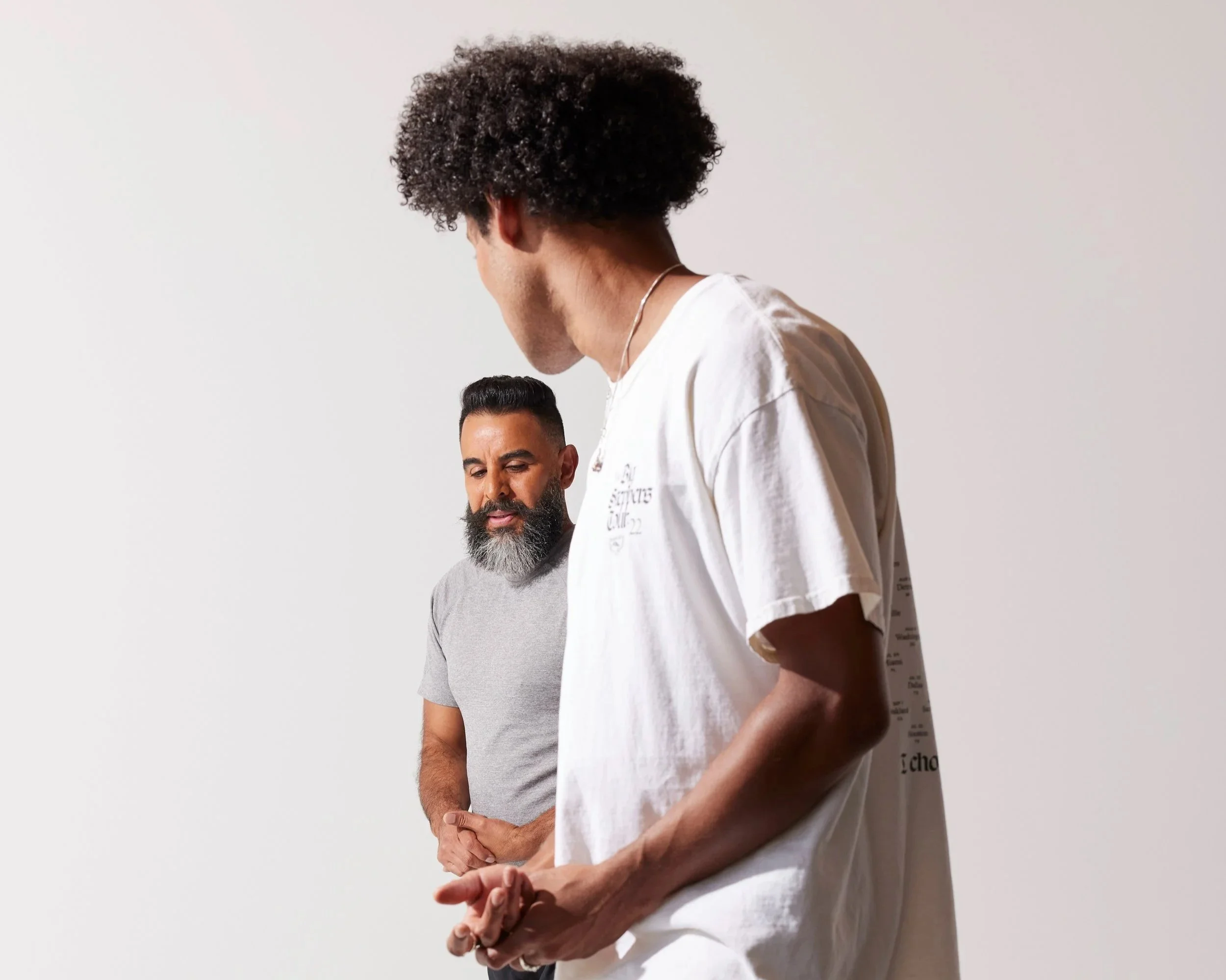 Three men standing against a plain, light-colored background, with one prominently in the foreground and two in the background, all with serious or contemplative expressions.