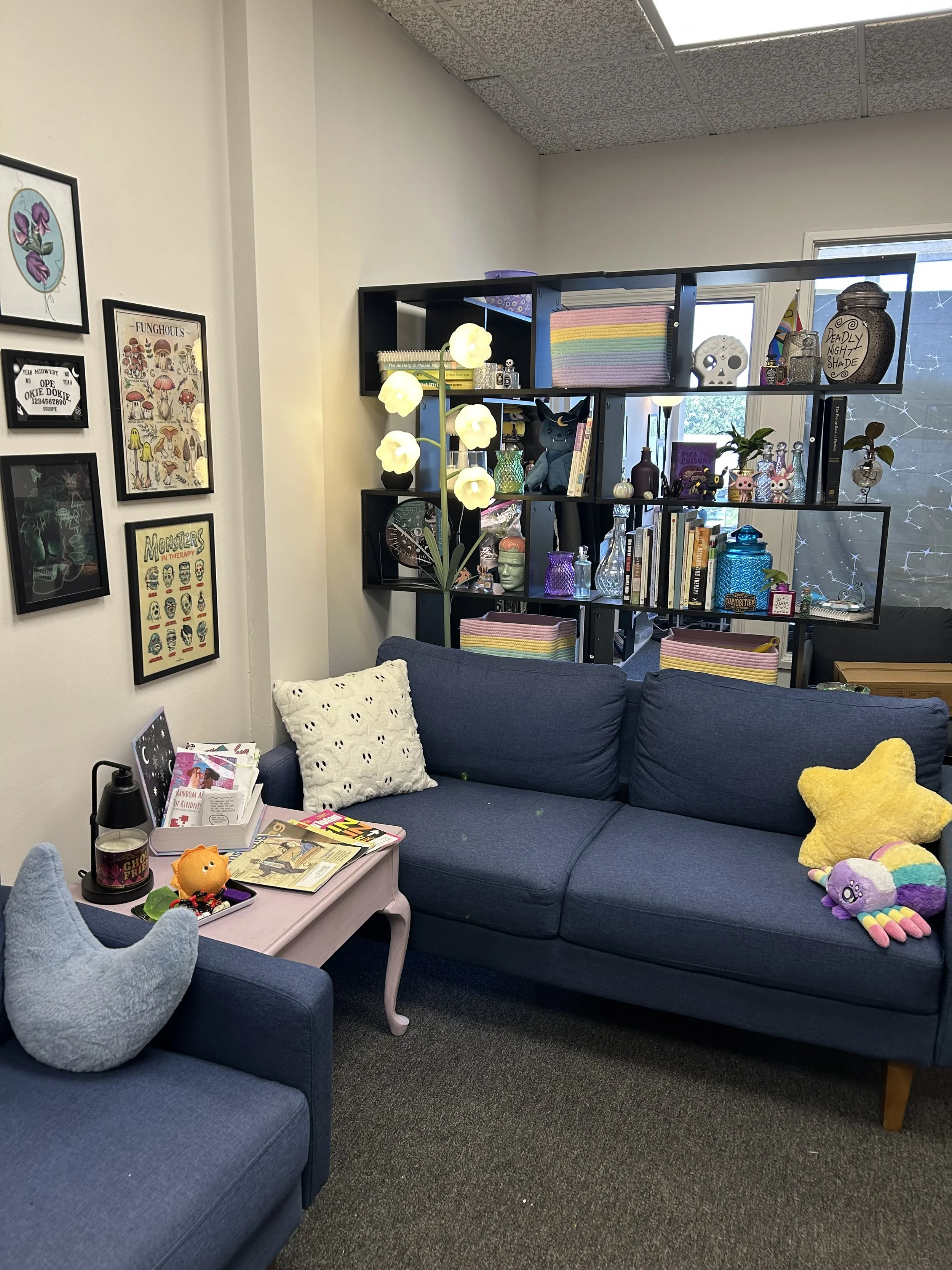 A cozy room with a blue sectional sofa and a pink coffee table. The sofa has plush toys, including a star-shaped pillow and a rainbow-colored plush. A black bookshelf with various books, decorations, glass bottles, and plants is behind the sofa. To the left, a blue armchair with a plush dinosaur toy is visible. The wall has framed artwork, and a unique lamp with multiple glowing flowers is in the room. The space has a mix of decorative and everyday items, creating a welcoming atmosphere.