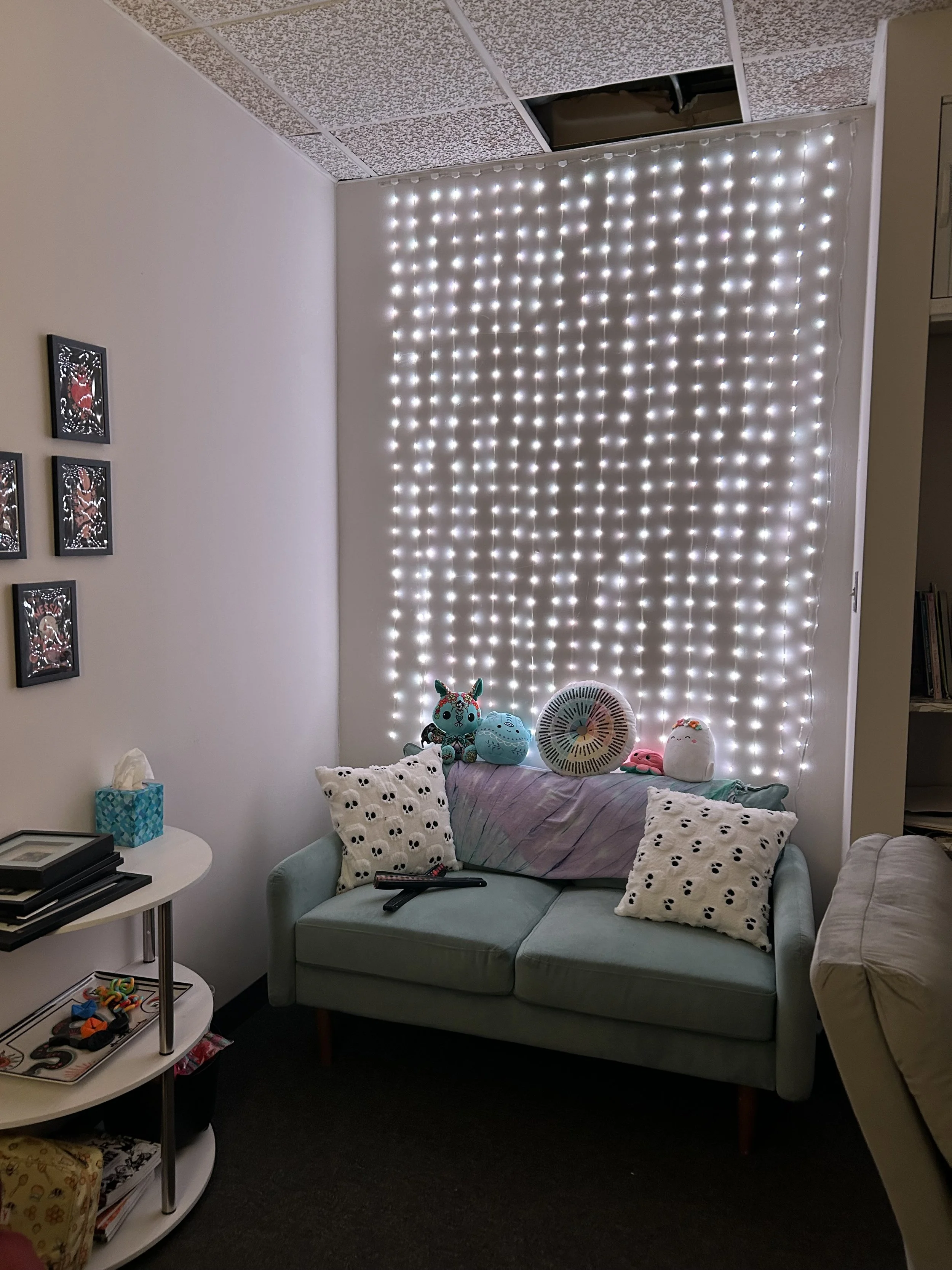 A room corner featuring a light green loveseat with pillow cushions and plush toys on top, illuminated by a grid of white LED strip lights on the wall behind. To the left, a white round side table holds a tissue box, books, and a remote. The wall has a small gallery of framed decorative cards.