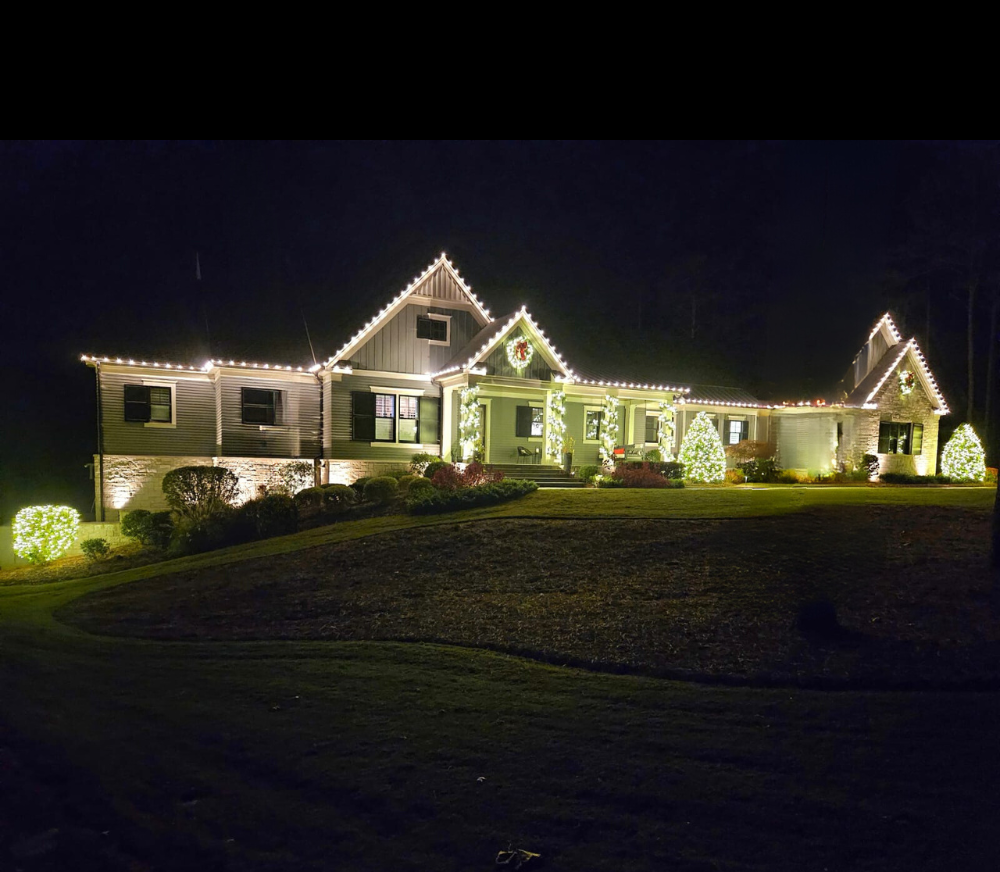 A large house decorated with festive holiday lights and wreaths, illuminated at night with a well-maintained lawn and bushes in front.