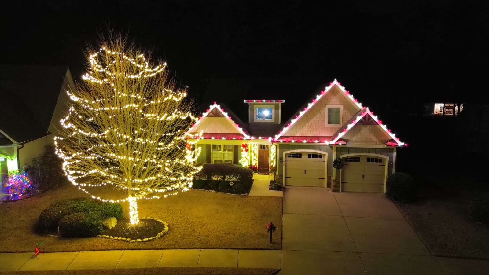 A house decorated for Christmas with colorful lights outlining the roof, a large lit-up tree in the front yard, and Christmas wreaths on the garage doors at night.