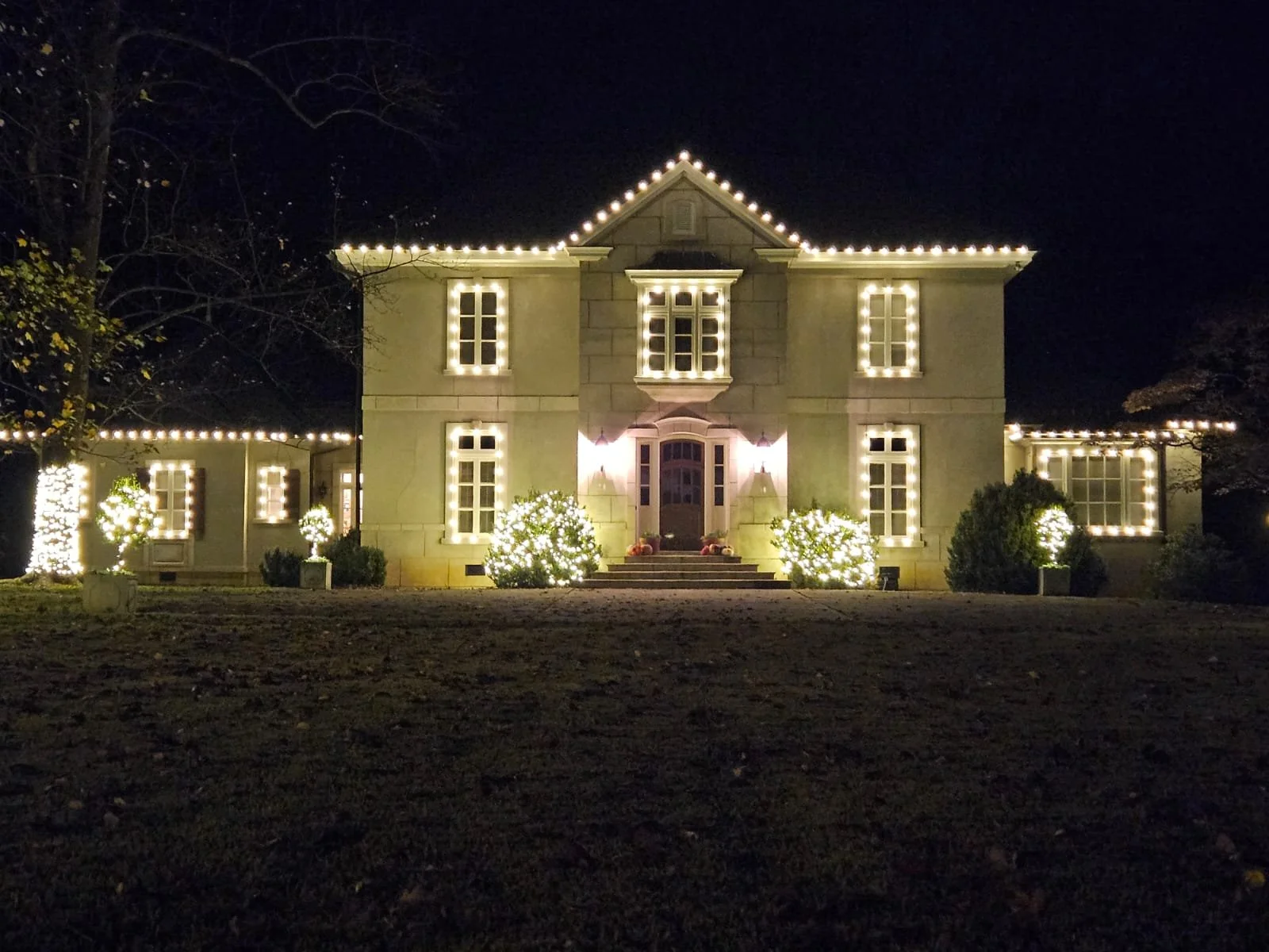 A large house decorated with string lights around the roofline, windows, and front porch at night. The house has a central entrance with steps leading up to the door, illuminated by two outdoor wall lights. Bushes with illuminated lights are on eithe