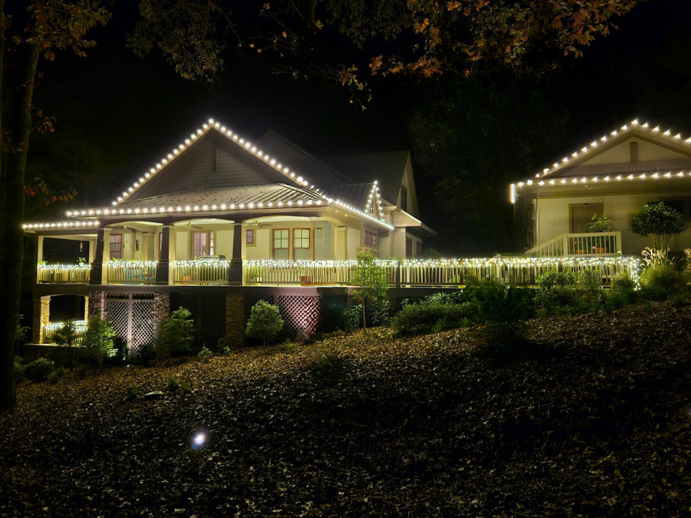 A house decorated with white string lights outlining the roof, porch, and railings at night, surrounded by trees and bushes.