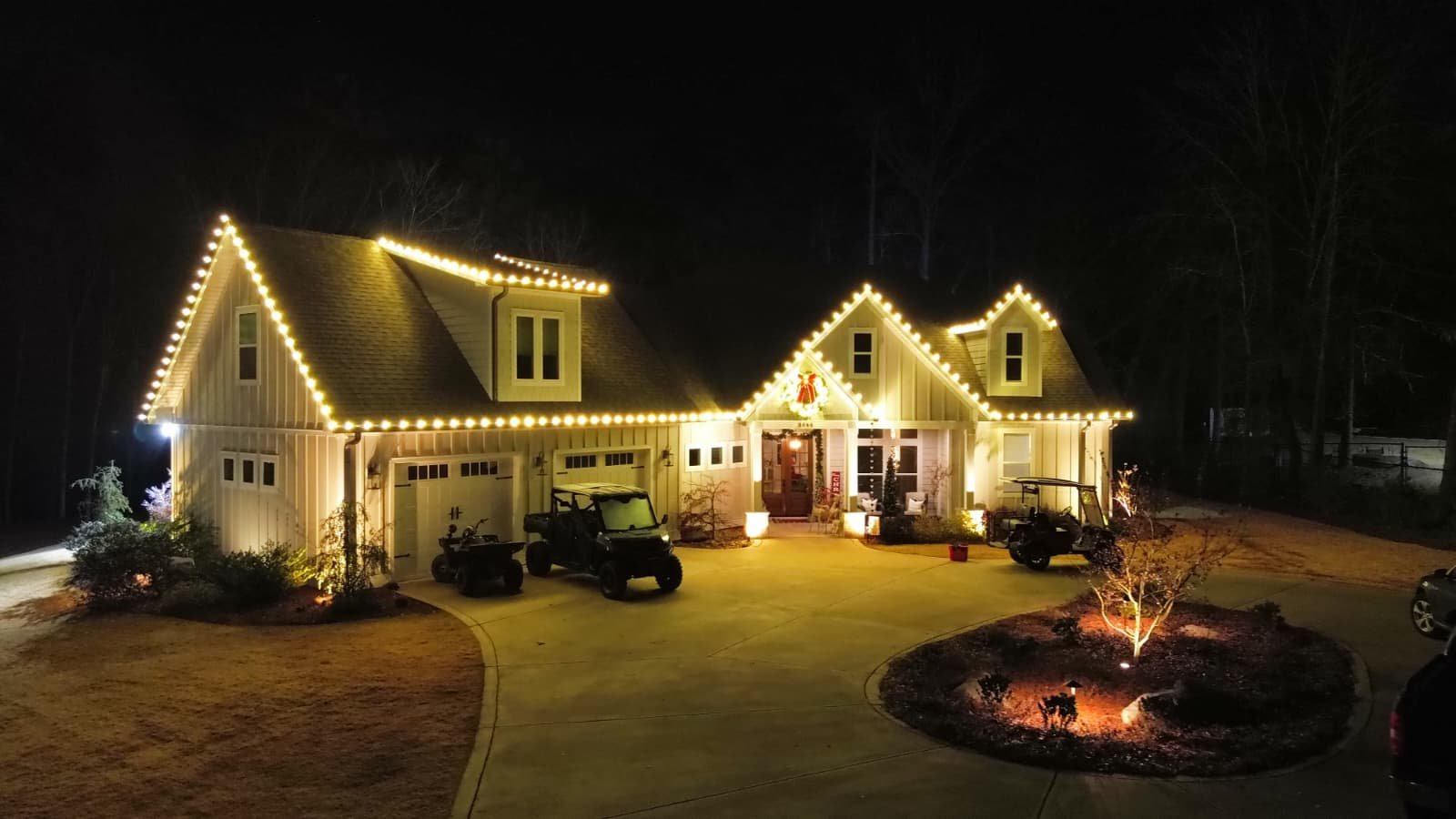 A large house decorated with string lights outlining the roof at night, with a landscaped front yard, driveway with parked vehicles and golf carts, and lit trees and shrubs.