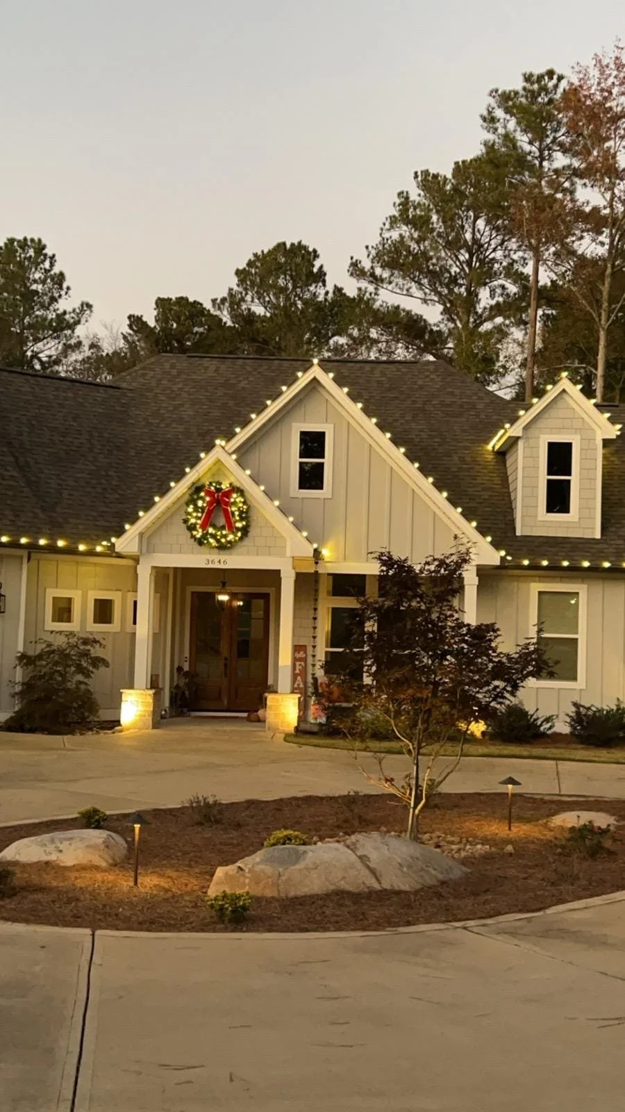 A house decorated with Christmas lights and a wreath with a red bow, set in a landscaped yard with rocks and small plants, during dusk.