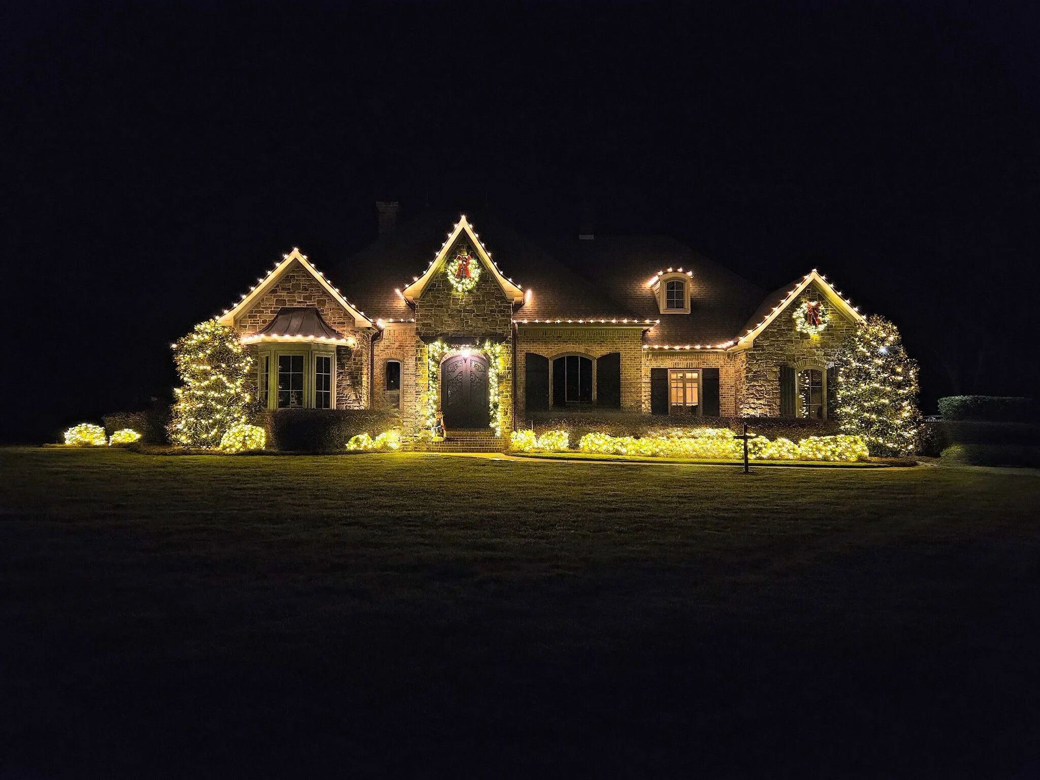 A large house decorated with Christmas lights and wreaths, with trees and bushes illuminated in front, under a dark night sky.