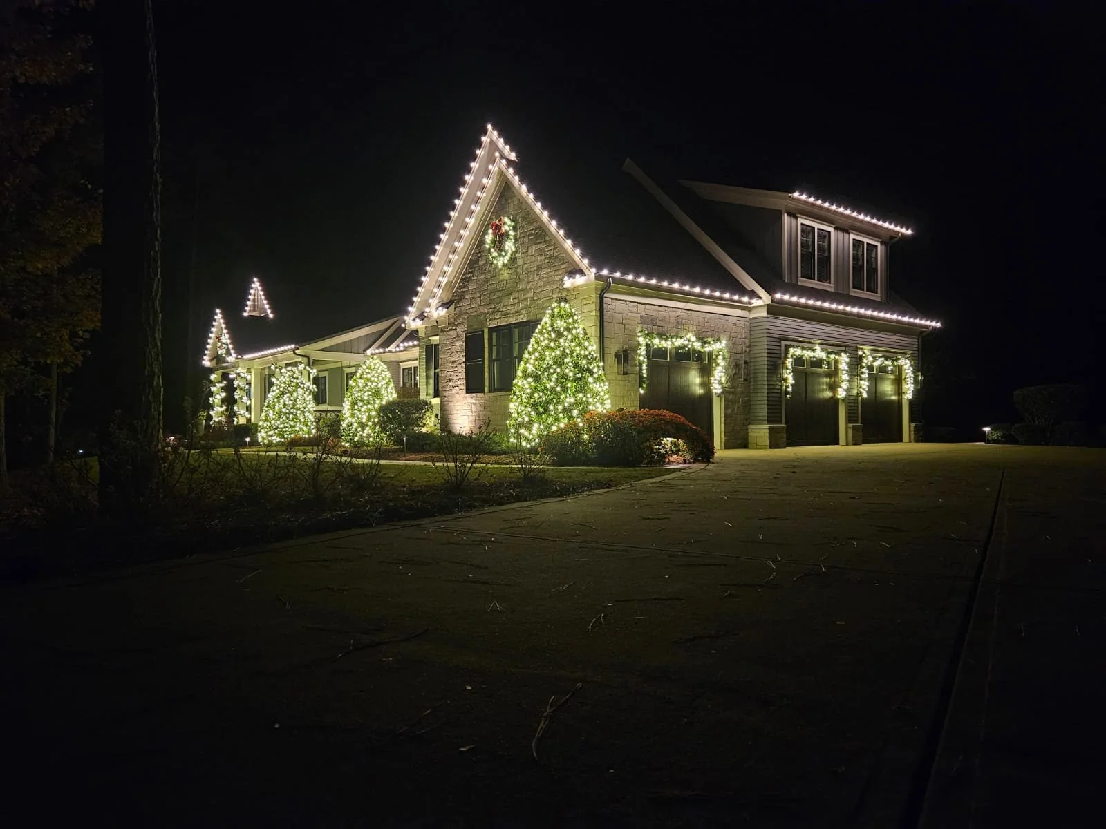 A house decorated with Christmas lights at night, featuring illuminated trees, wreaths, and outline lighting on the roof and garage.