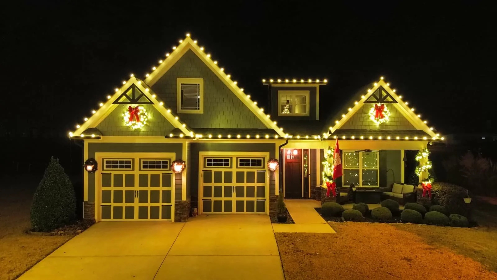 A house decorated for Christmas with yellow lights outlining the roof, wreaths with red bows, and a red flag on the porch at night.