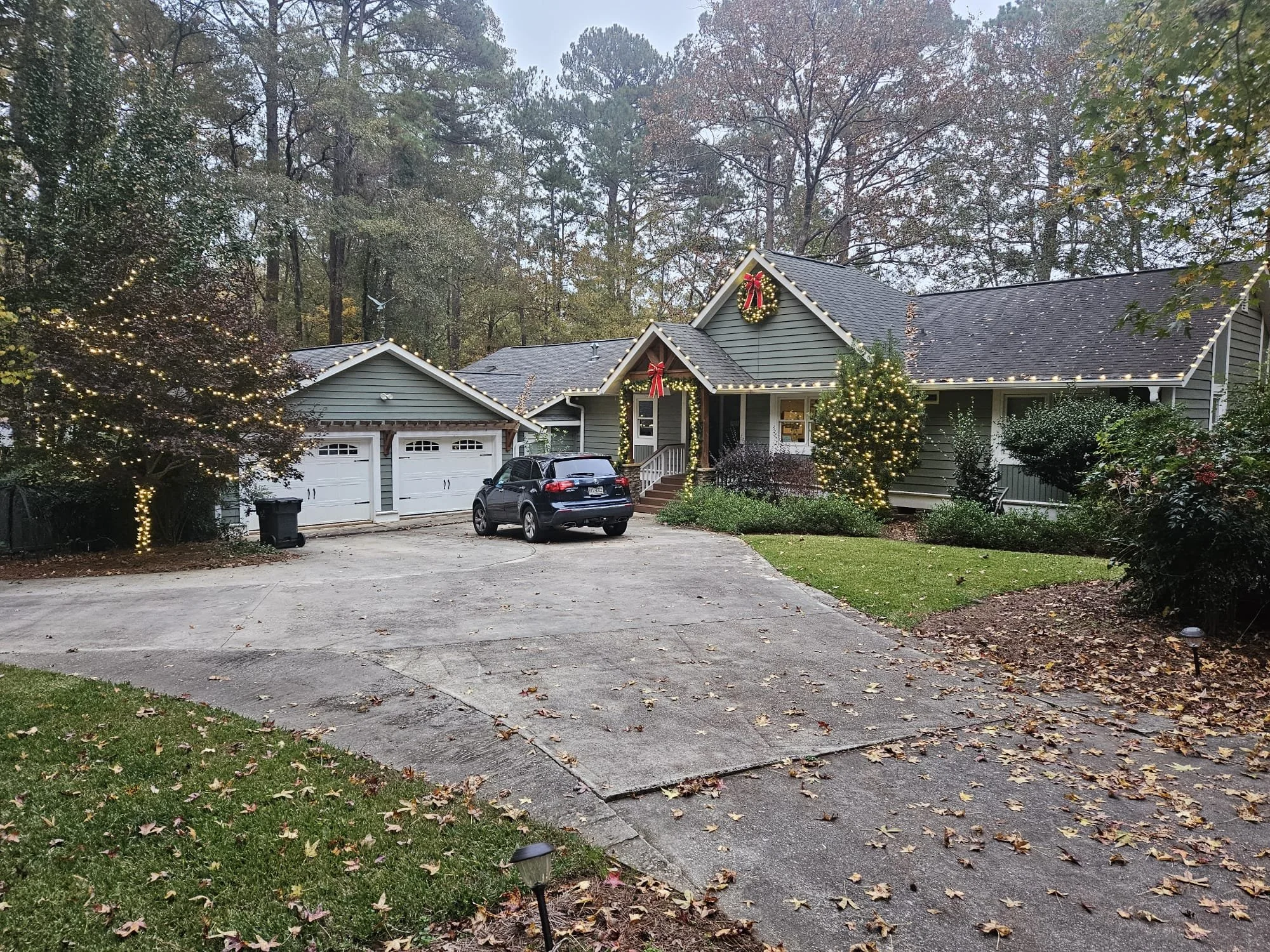 A house decorated for Christmas with wreaths and string lights. The house has a front porch, an attached garage, and is surrounded by trees with holiday decorations. A car is parked in the driveway, and there is a lawn with fallen leaves.