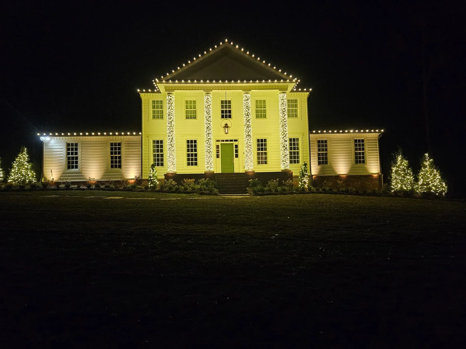 A large house decorated with Christmas lights at night. The house is painted light yellow, with bright white string lights outlining the roof and columns. The front yard has lit Christmas trees and small bushes.