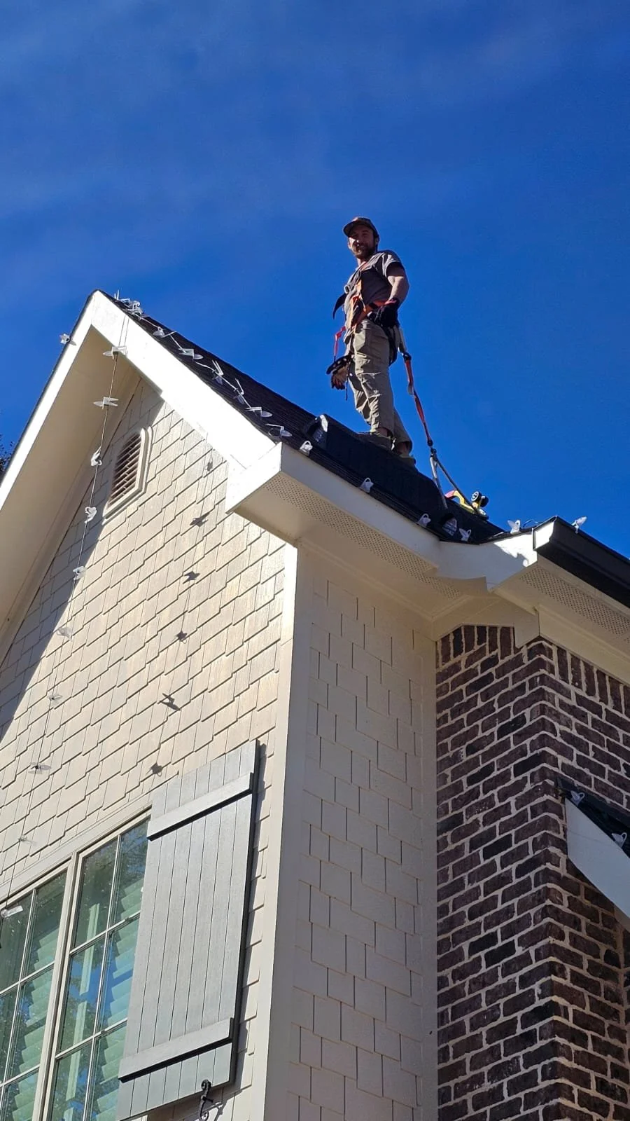 A man standing on the roof of a house with a safety harness, against a blue sky.