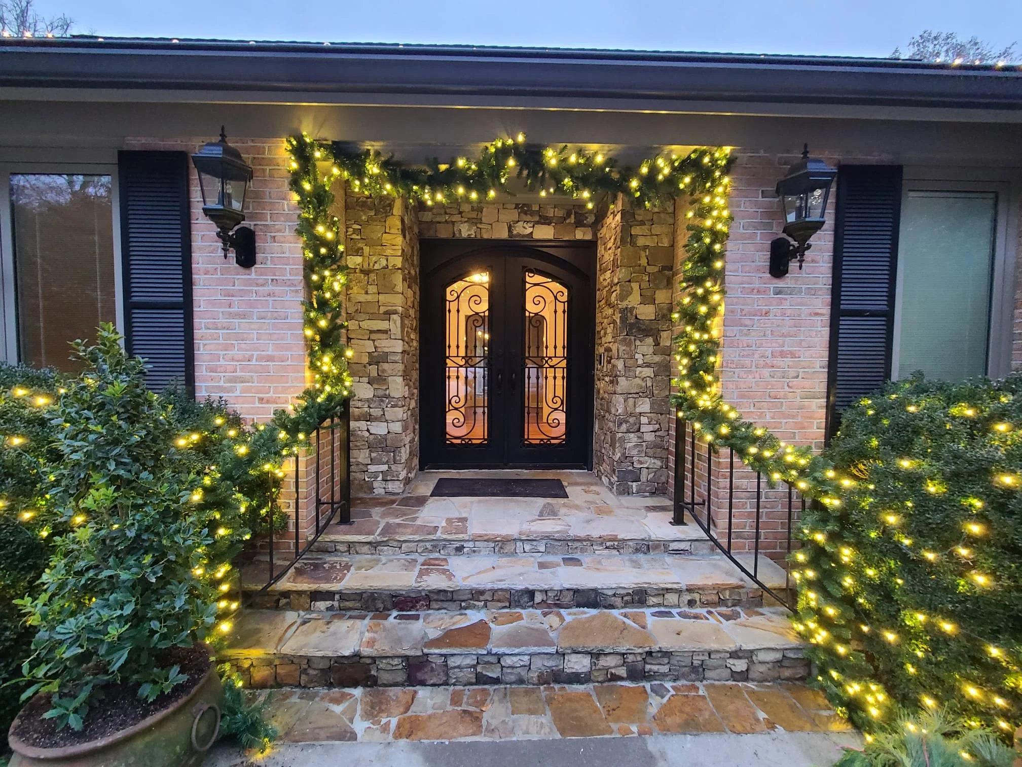 Front porch of a house decorated for Christmas with yellow string lights, greenery, and black lantern-style exterior lights. The porch has brick and stone walls and a wrought iron door.